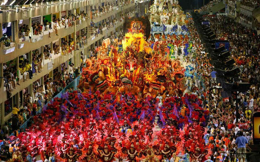 Carnival 桌布 壁紙 — 一場耀眼的夜間嘉年華遊行，充滿活力的紅色舞者和高聳的金色花車，擠滿了兩旁陽台觀眾的街道。