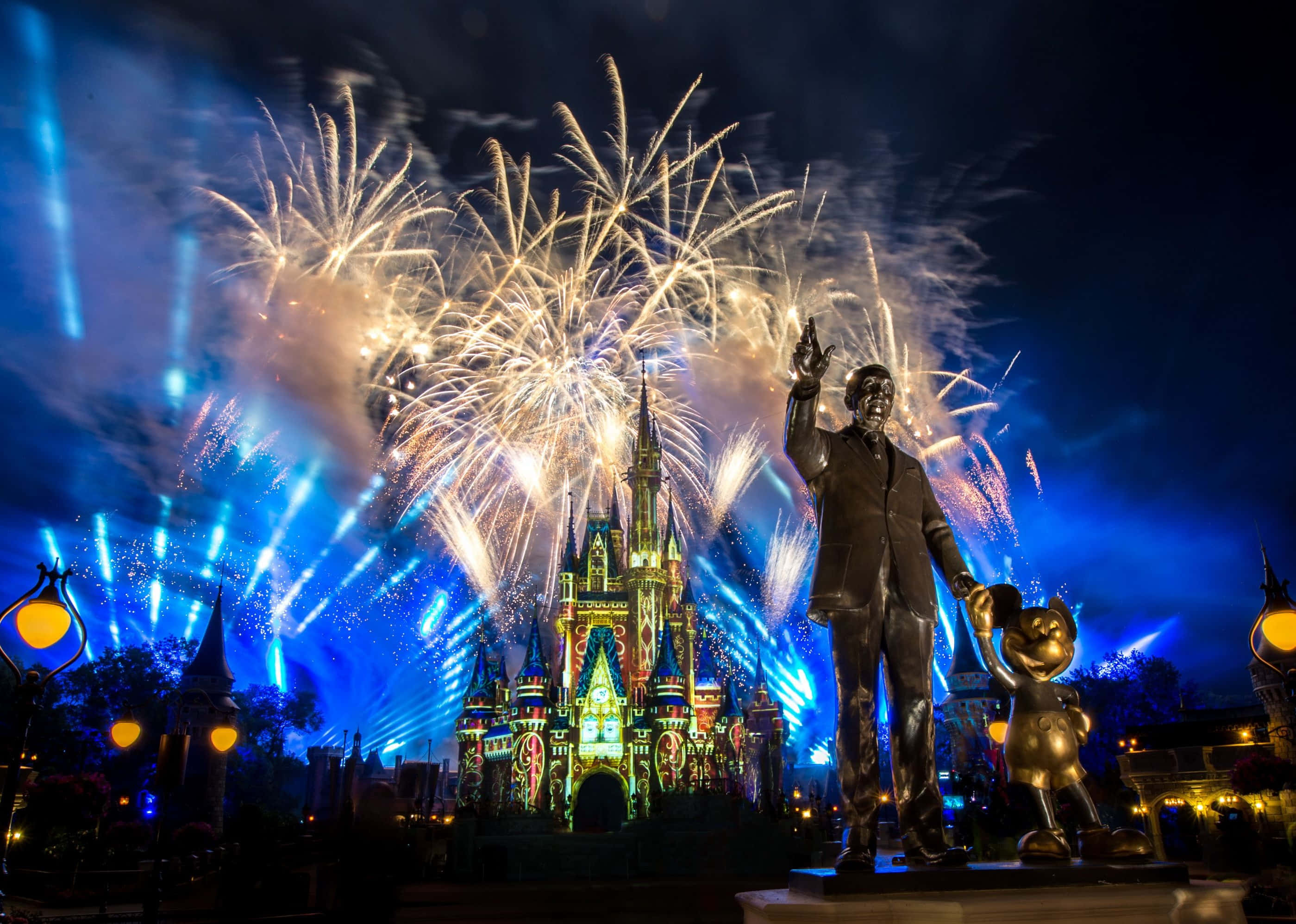 Cool Disney 桌布 壁紙 — 一場耀眼的夜間煙火表演在燈火通明的城堡上空綻放，前景是華特迪士尼和米奇老鼠的雕像。