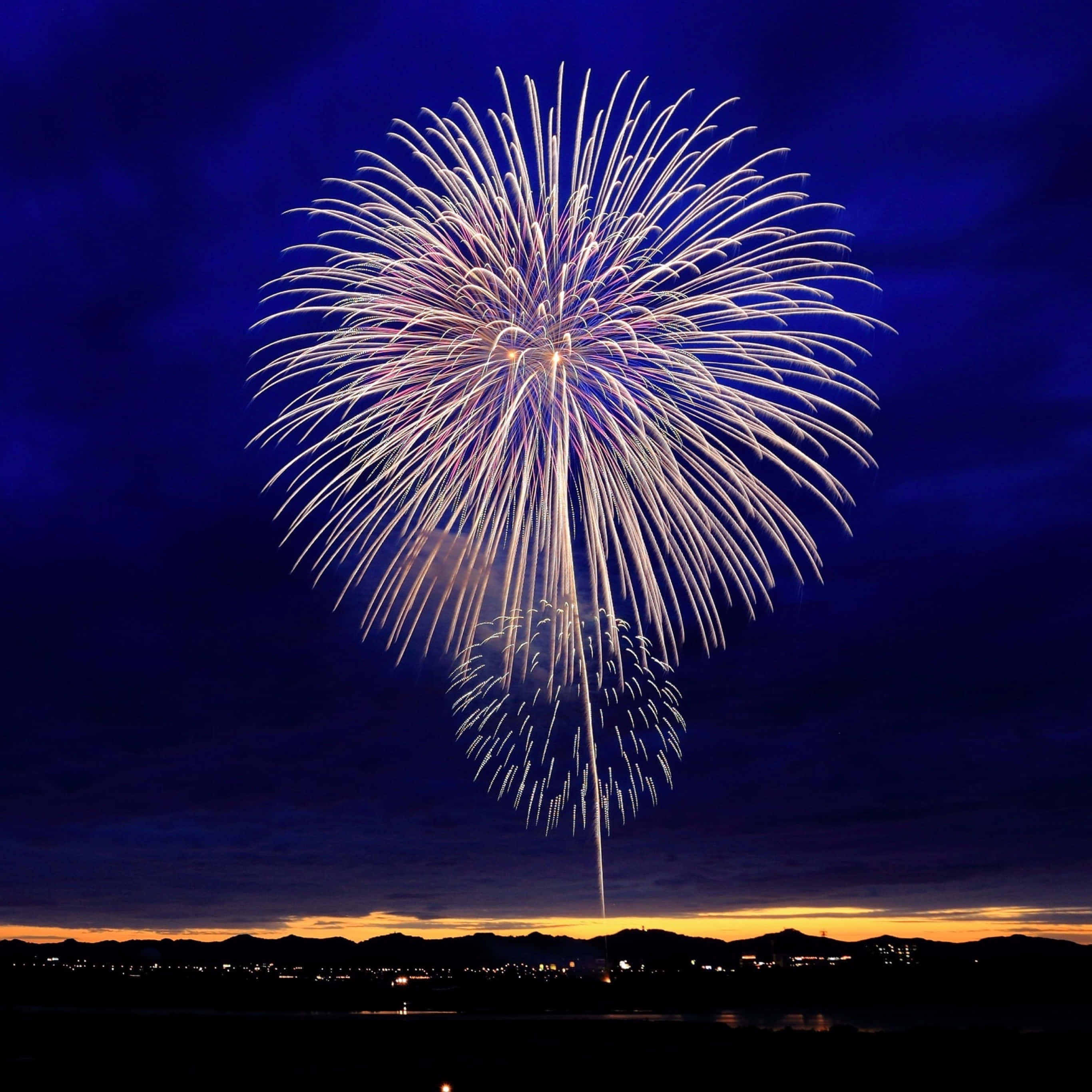 firework 桌布 壁紙 — 一場耀眼的白色和紫色煙火爆炸在深藍色夜空綻放，映襯著剪影般的山脈和下方閃爍的城市燈光。