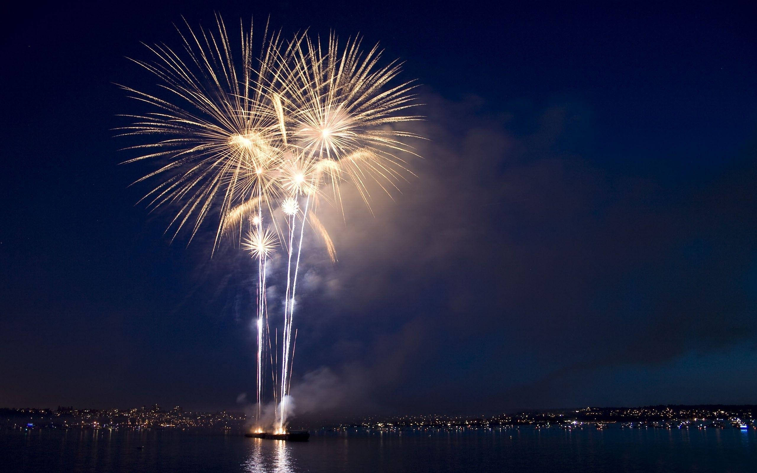 Fireworks 桌布 壁紙 — 一場耀眼的金色煙火表演在深色水面上爆炸，遠處的城市燈光在深邃的夜空下閃爍。