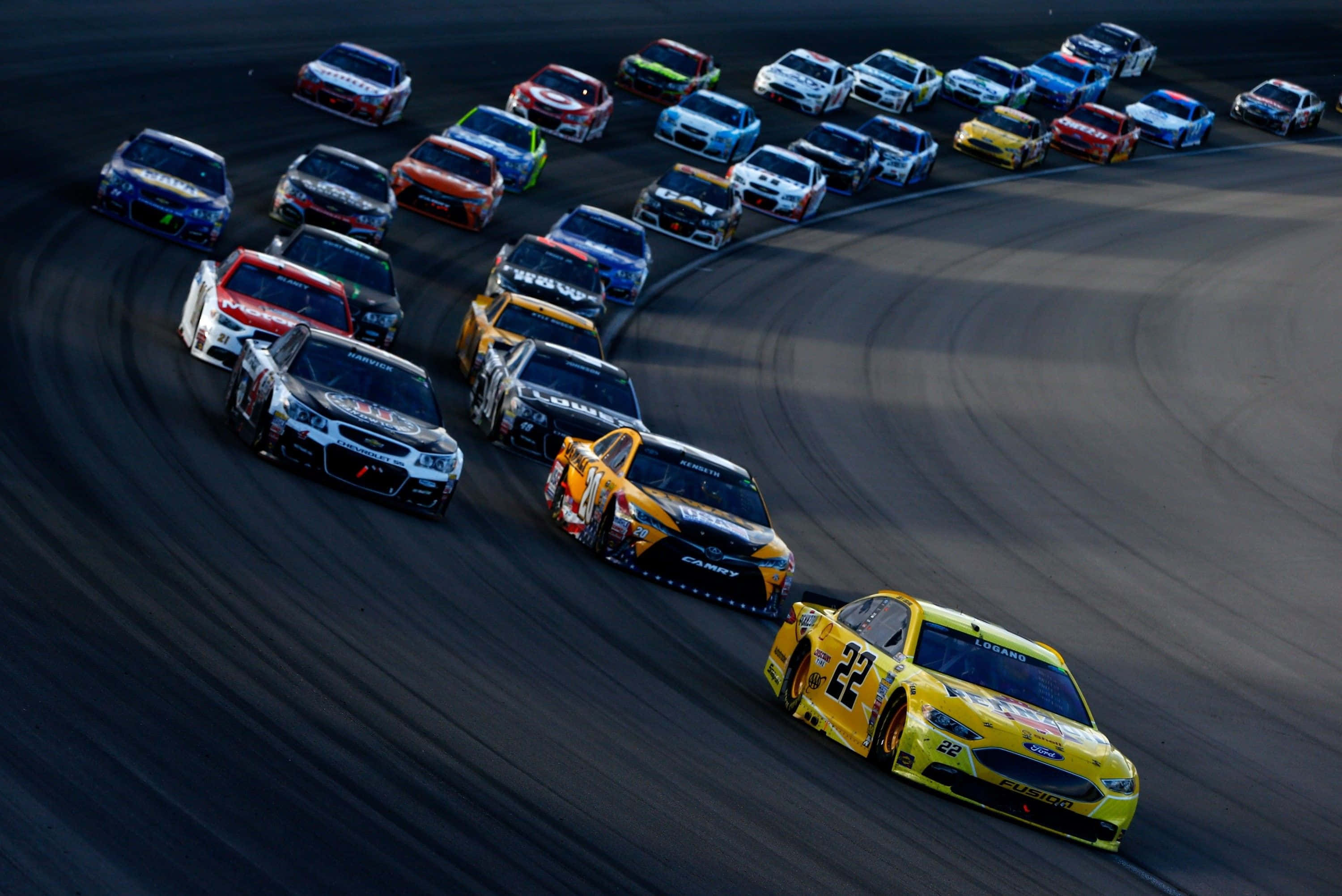 nascar 桌布 壁紙 — 一場高速NASCAR比賽，數十輛色彩鮮豔的賽車緊密排列，在深色瀝青賽道上駛過一個大彎。