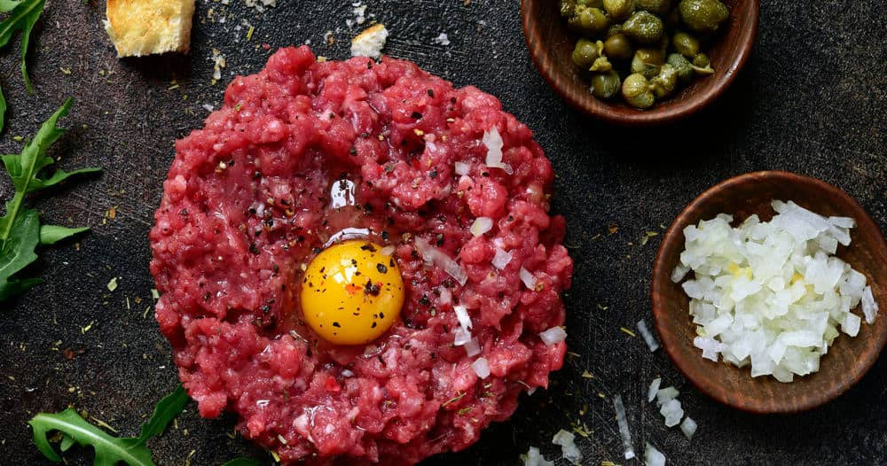 meat 桌布 壁紙 — 一塊充滿活力的生牛肉他他，中央放著亮黃色的蛋黃，周圍環繞著酸豆、洋蔥和芝麻菜，置於深色質樸的表面上。