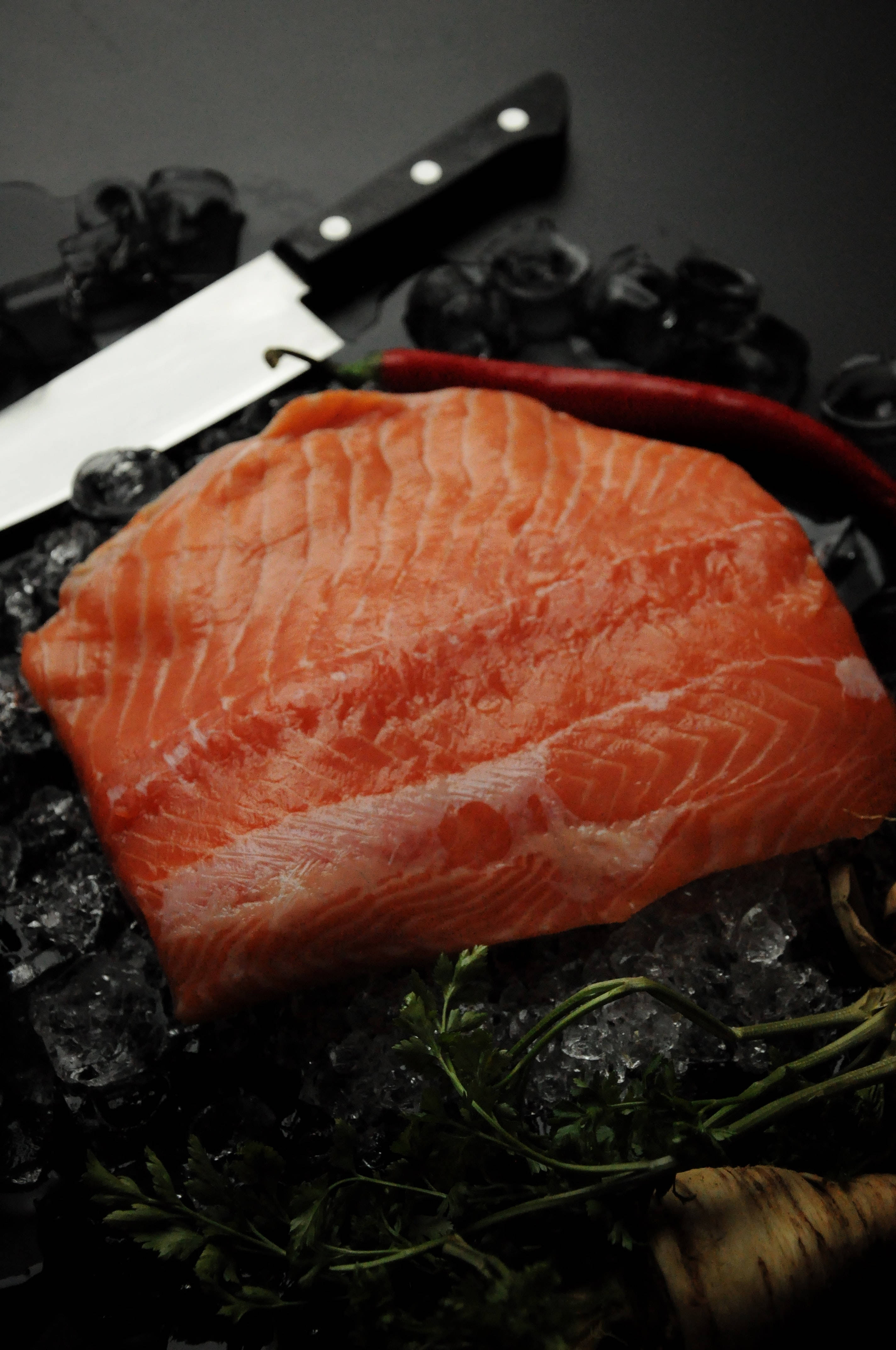 魚 桌布 壁紙 — 一塊充滿活力的生鮭魚片放在碎冰上，旁邊有刀、辣椒和香草，在戲劇性的昏暗燈光下。