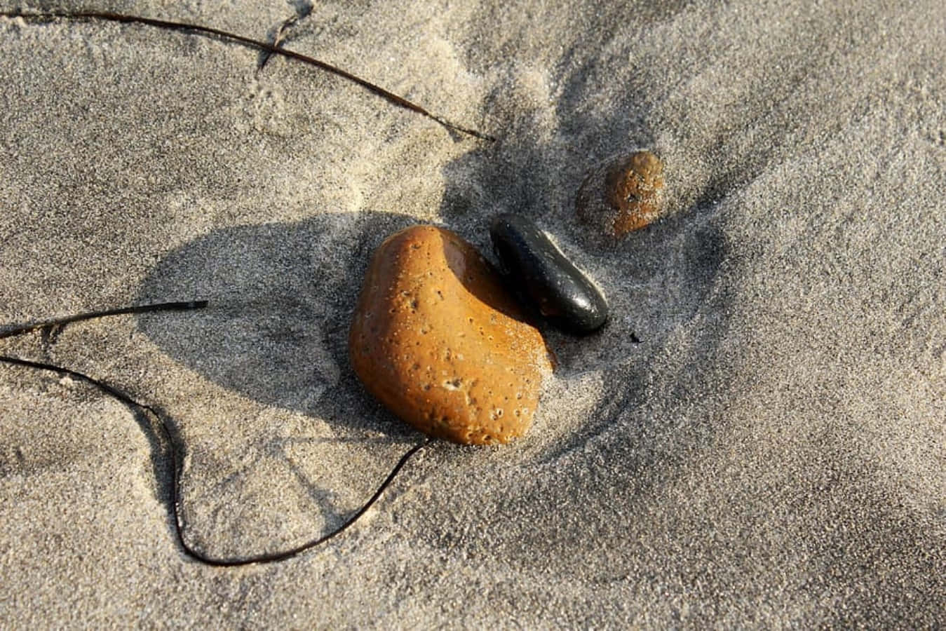 Stone 桌布 壁紙 — 一塊光滑的橙色石頭和深色鵝卵石，在溫暖的陽光下，躺在佈滿海藻的紋理沙子上。