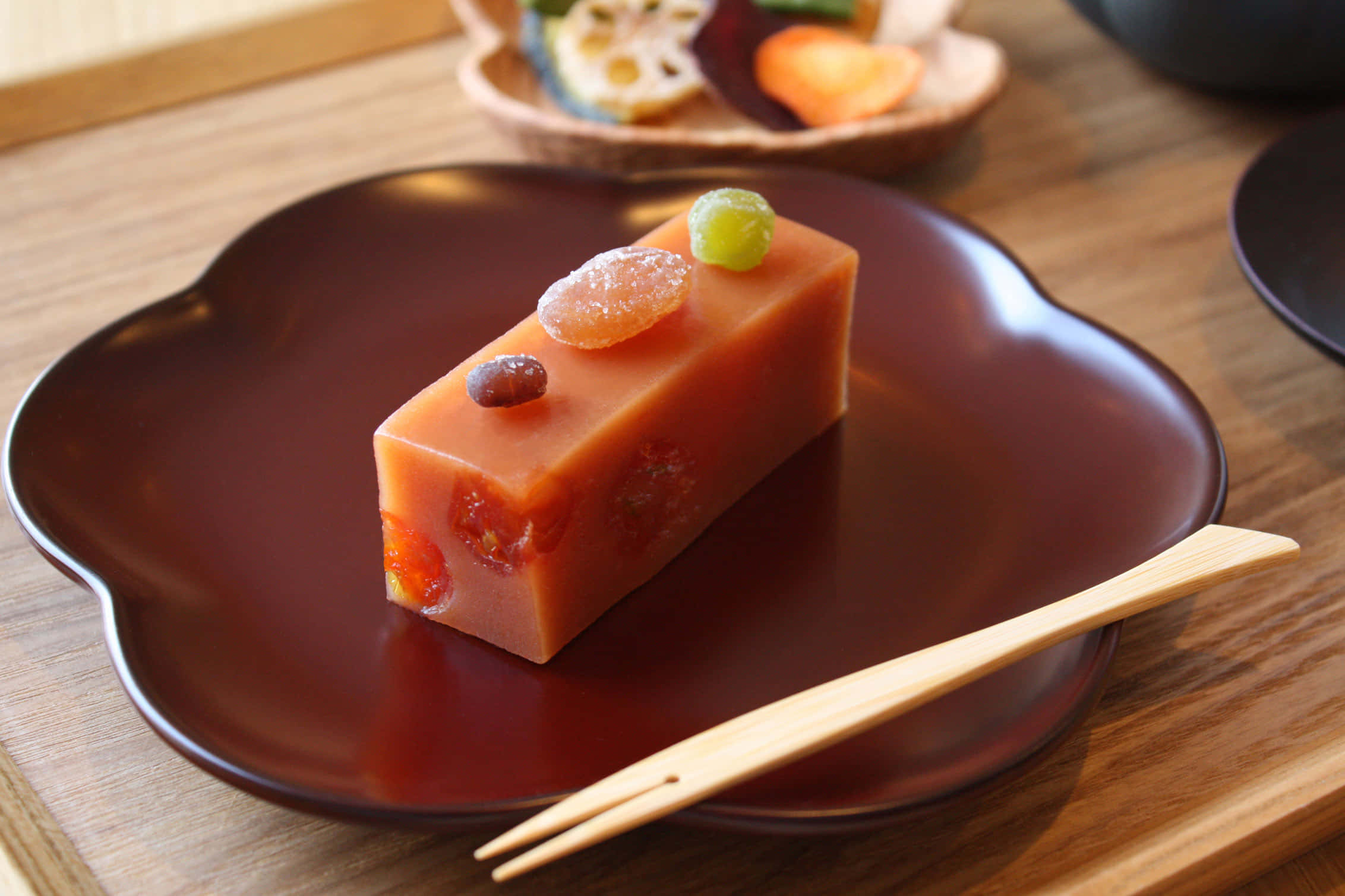 Japanese Sweets 桌布 壁紙 — 一塊半透明橙色日式麻糬甜點的長方形切片，上面放有蜜餞水果，置於深棕色花形盤子上，旁邊有木筷子。