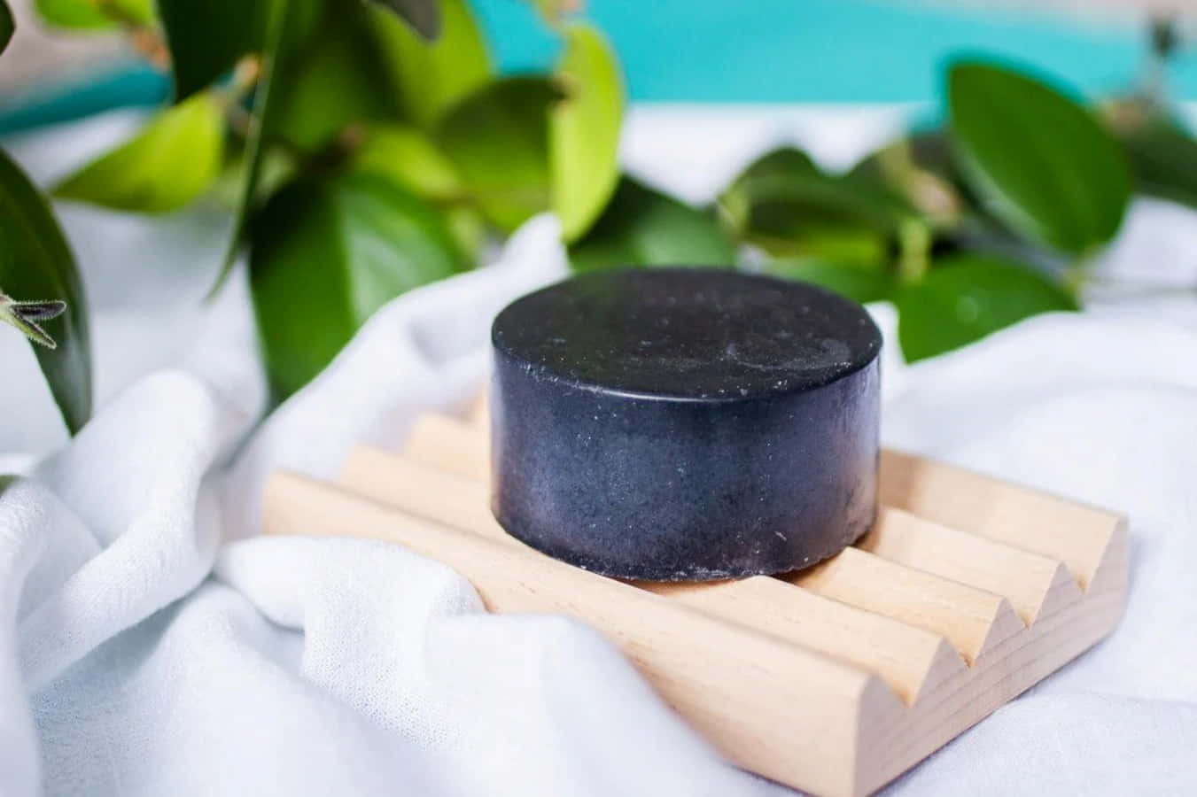 Black Soap 桌布 壁紙 — 一塊啞光黑色圓柱形肥皂放在淺色木條碟上，碟下有一塊白布，背景有柔焦的綠葉。