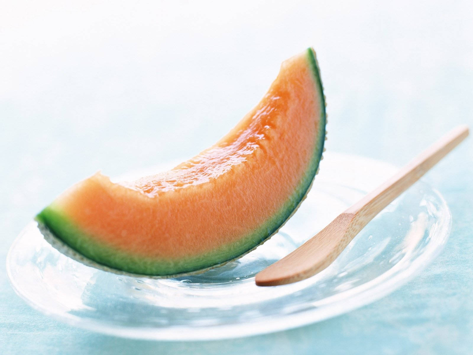 Fruit 桌布 壁紙 — 一塊多汁的哈密瓜片放在透明玻璃盤上，旁邊有木湯匙，背景是柔和的藍色。