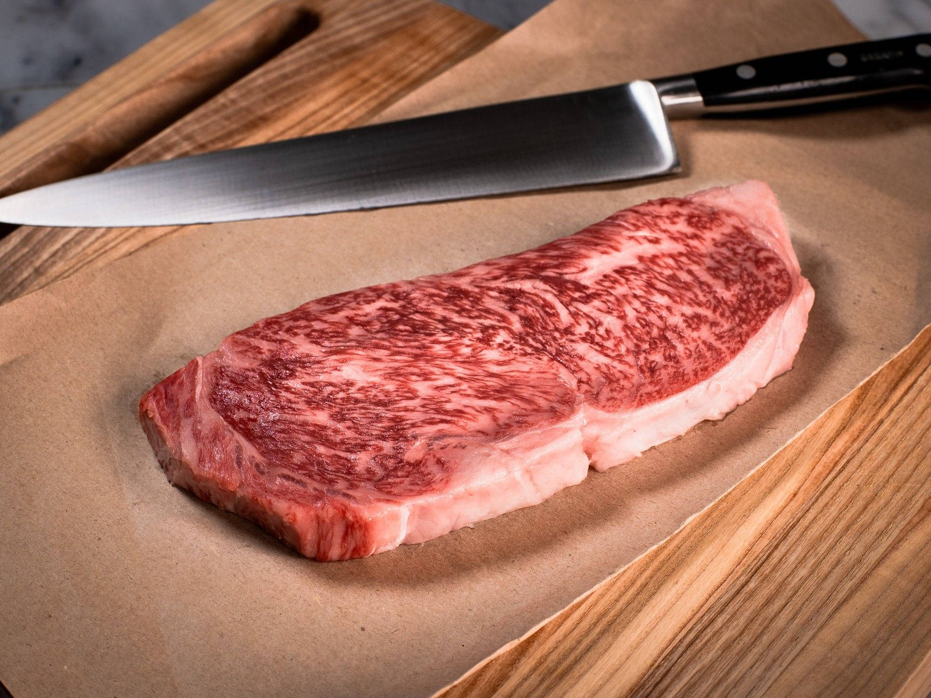 meat 桌布 壁紙 — 一塊多汁的大理石紋牛排放在木砧板上的棕色紙上，旁邊有一把廚師刀。