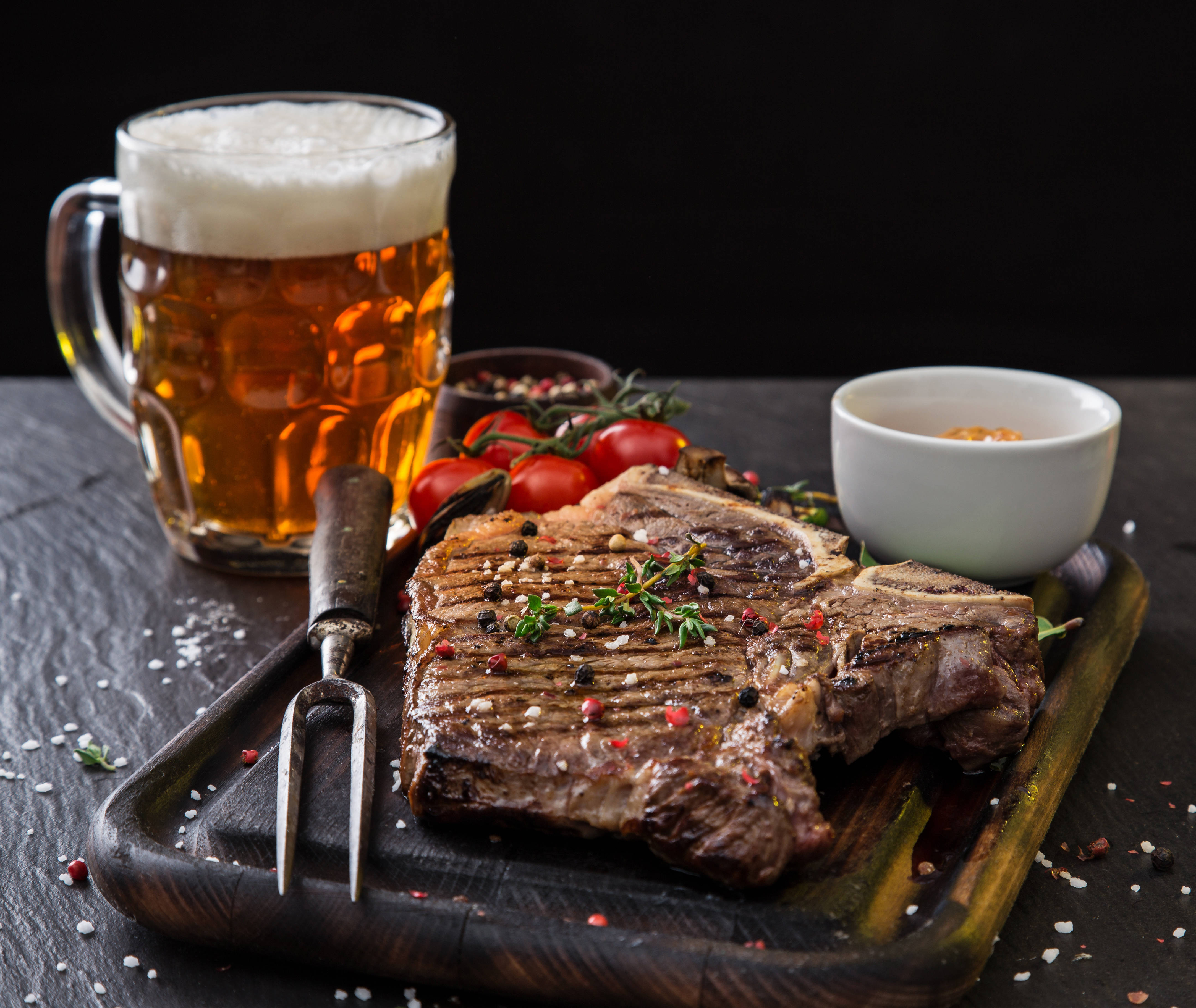 meat 桌布 壁紙 — 一塊多汁的烤T骨牛排，用香草和香料調味，放在木板上，旁邊有一杯泡沫啤酒和櫻桃番茄。