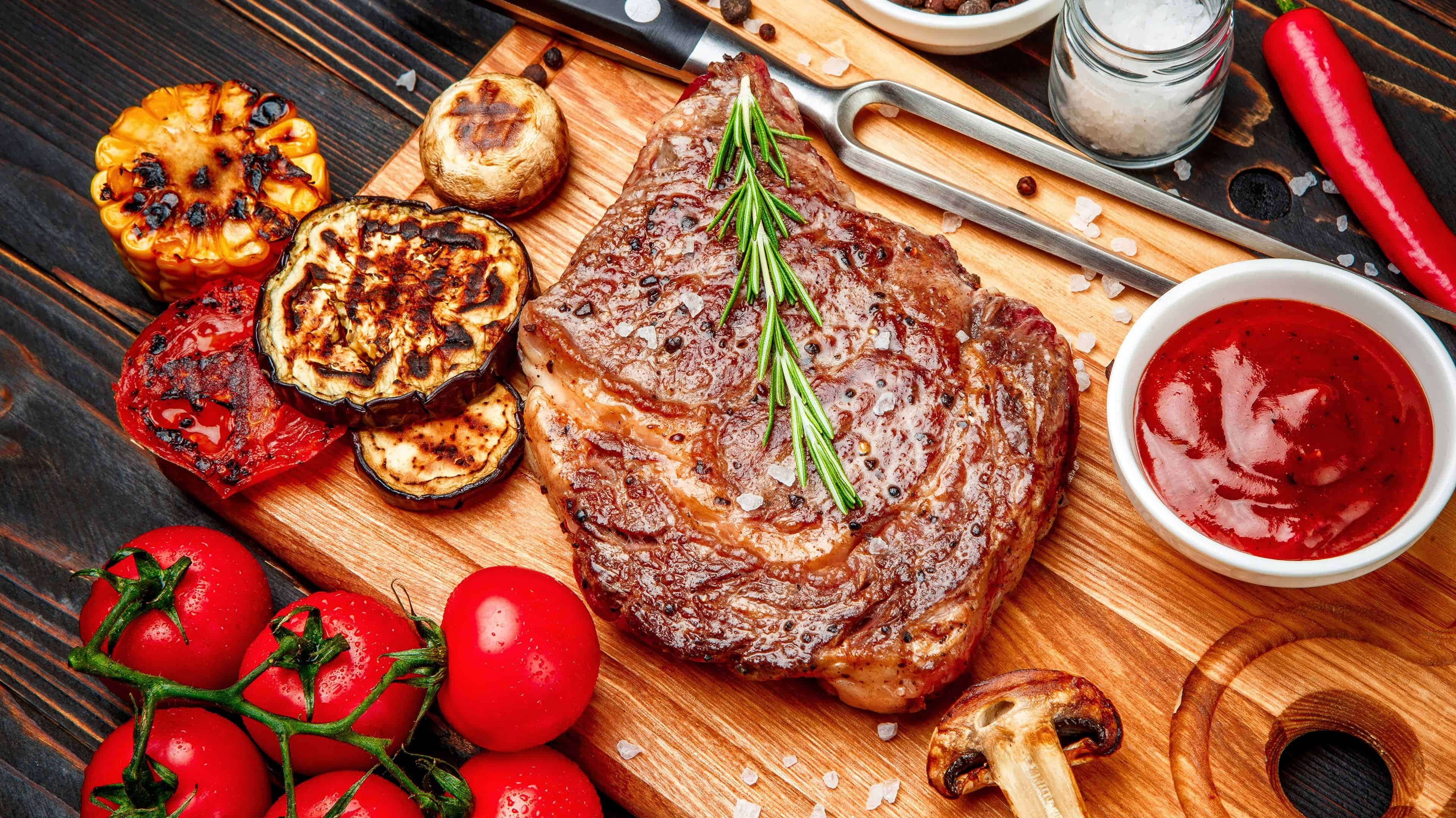meat 桌布 壁紙 — 一塊多汁的烤牛排，飾以迷迭香，放在木板上，周圍是烤蔬菜和新鮮番茄。
