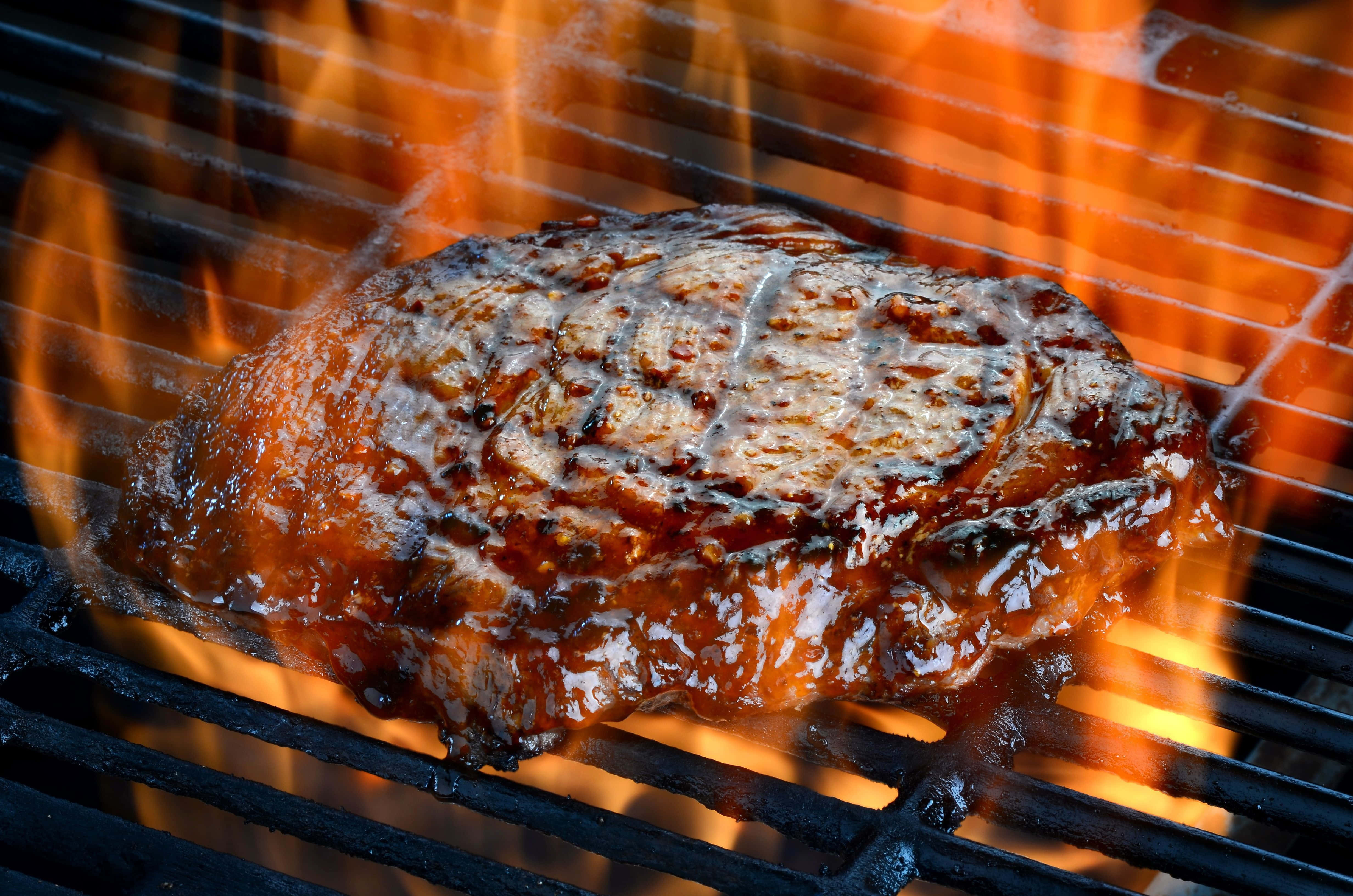 Steak 桌布 壁紙 — 一塊多汁的牛排正在黑色烤架上於熊熊烈火中滋滋作響，帶有明顯的焦痕和閃亮的肉汁。