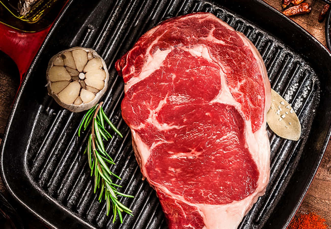 meat 桌布 壁紙 — 一塊多汁的生牛排放在黑色烤盤上，旁邊有大蒜、迷迭香和月桂葉，準備烹調。