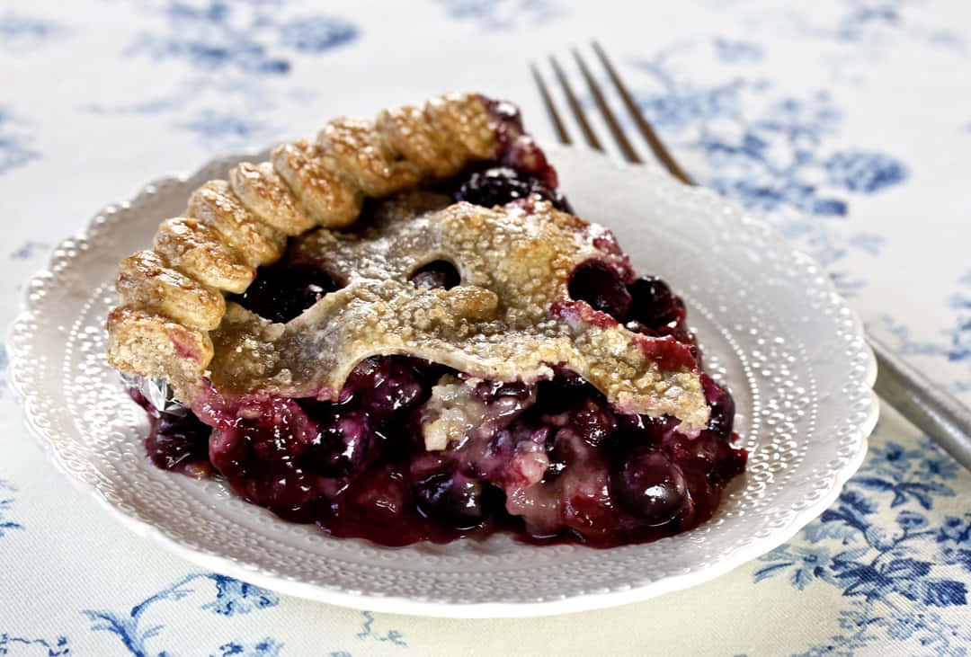 Blueberry Pie 桌布 壁紙 — 一塊多汁的藍莓派，帶有金黃酥脆的外皮，放在白色花紋盤上，旁邊有一把叉子。