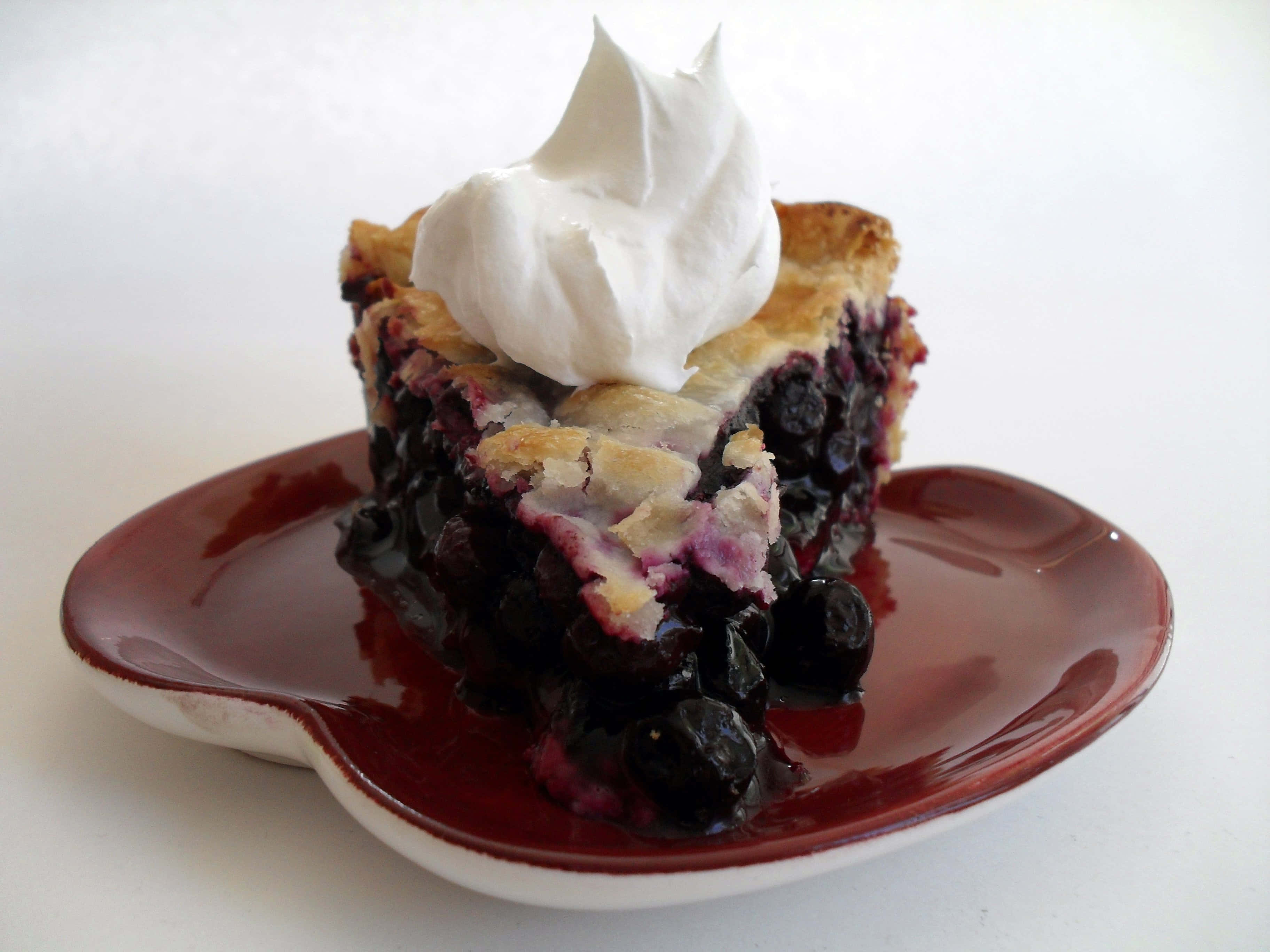 Blueberry Pie 桌布 壁紙 — 一塊多汁的藍莓派，派皮酥脆，配上鮮奶油，放在亮面紅色盤子上，背景為白色。