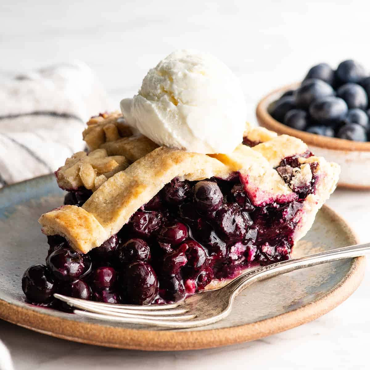 Blueberry Pie 桌布 壁紙 — 一塊多汁的藍莓派，配上酥脆的外皮和香草冰淇淋，放在一個鄉村風格的盤子上，旁邊有叉子和一碗新鮮莓果。