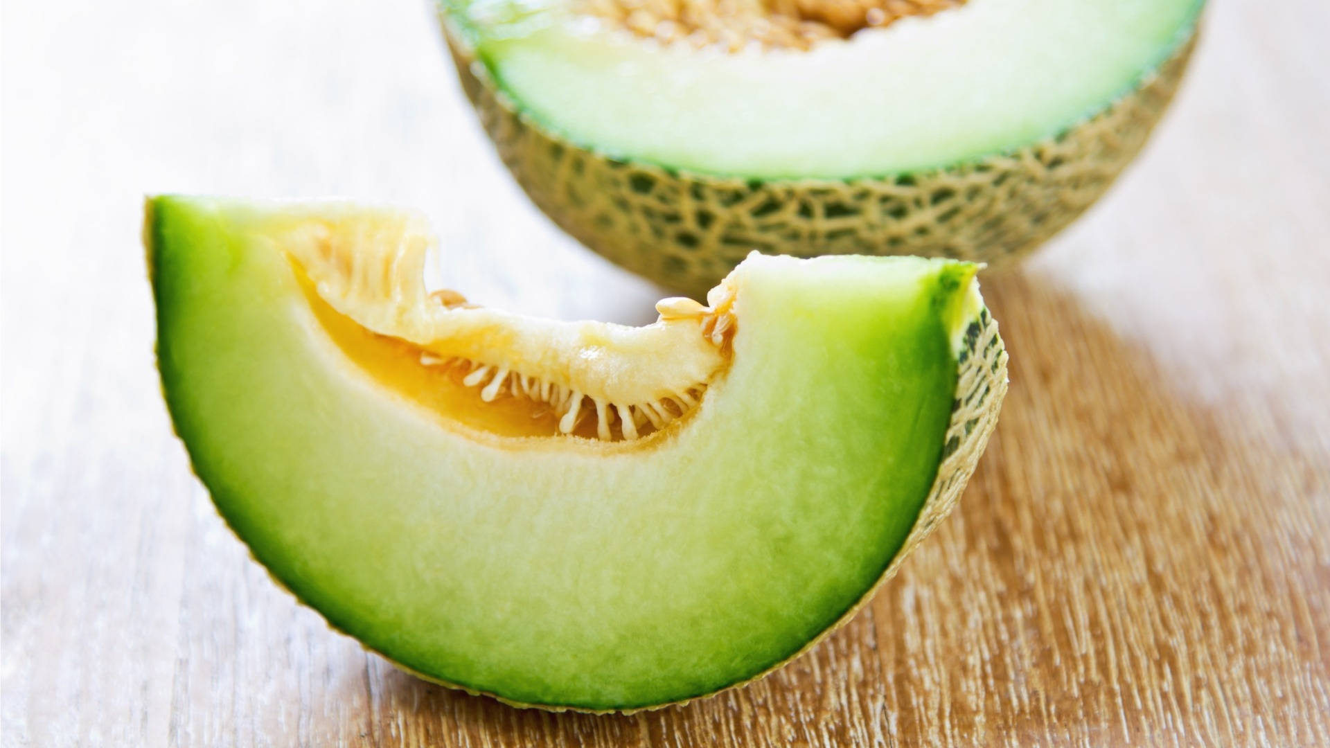 Fruits 桌布 壁紙 — 一塊多汁的蜜瓜片放在木質表面上，旁邊是一個切半的蜜瓜，露出淡綠色的果肉和種子。
