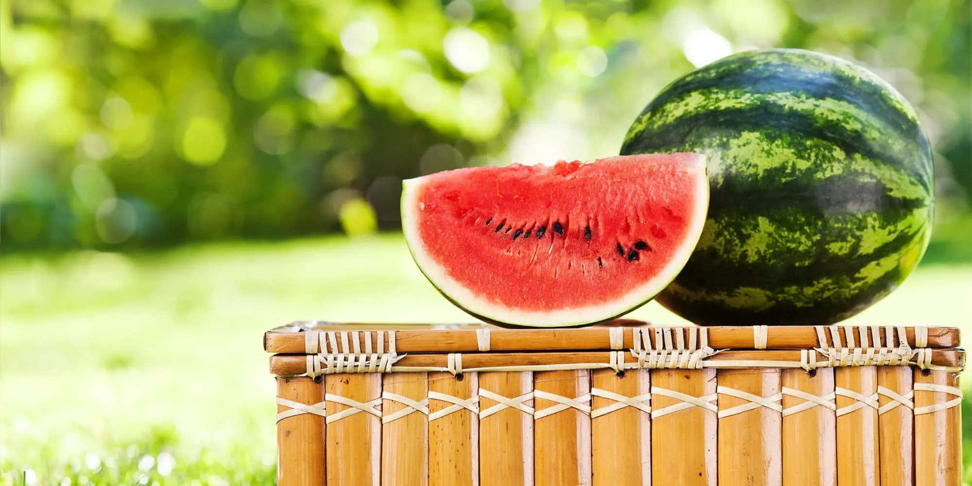 Watermelon 桌布 壁紙 — 一塊多汁的西瓜片和整個甜瓜放在一個編織竹野餐籃上，在陽光明媚的草地上。