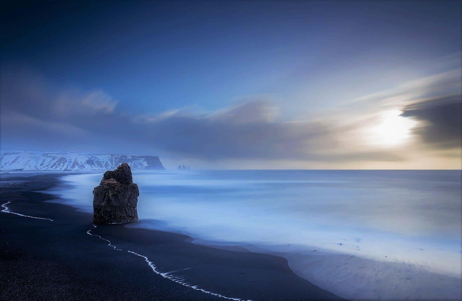Winter Beach 桌布 壁紙 — 一塊孤獨崎嶇的岩石矗立在黑色火山沙灘上，霧氣繚繞的海浪在戲劇性的暮色天空下流過。