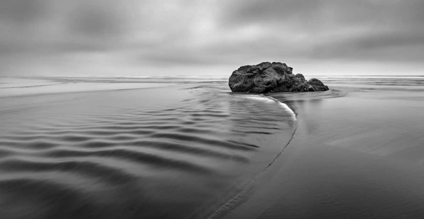 Black And White Ocean 桌布 壁紙 — 一塊孤獨的岩石矗立在霧氣瀰漫的黑白海灘場景中，水面平滑泛起漣漪，天空多雲。