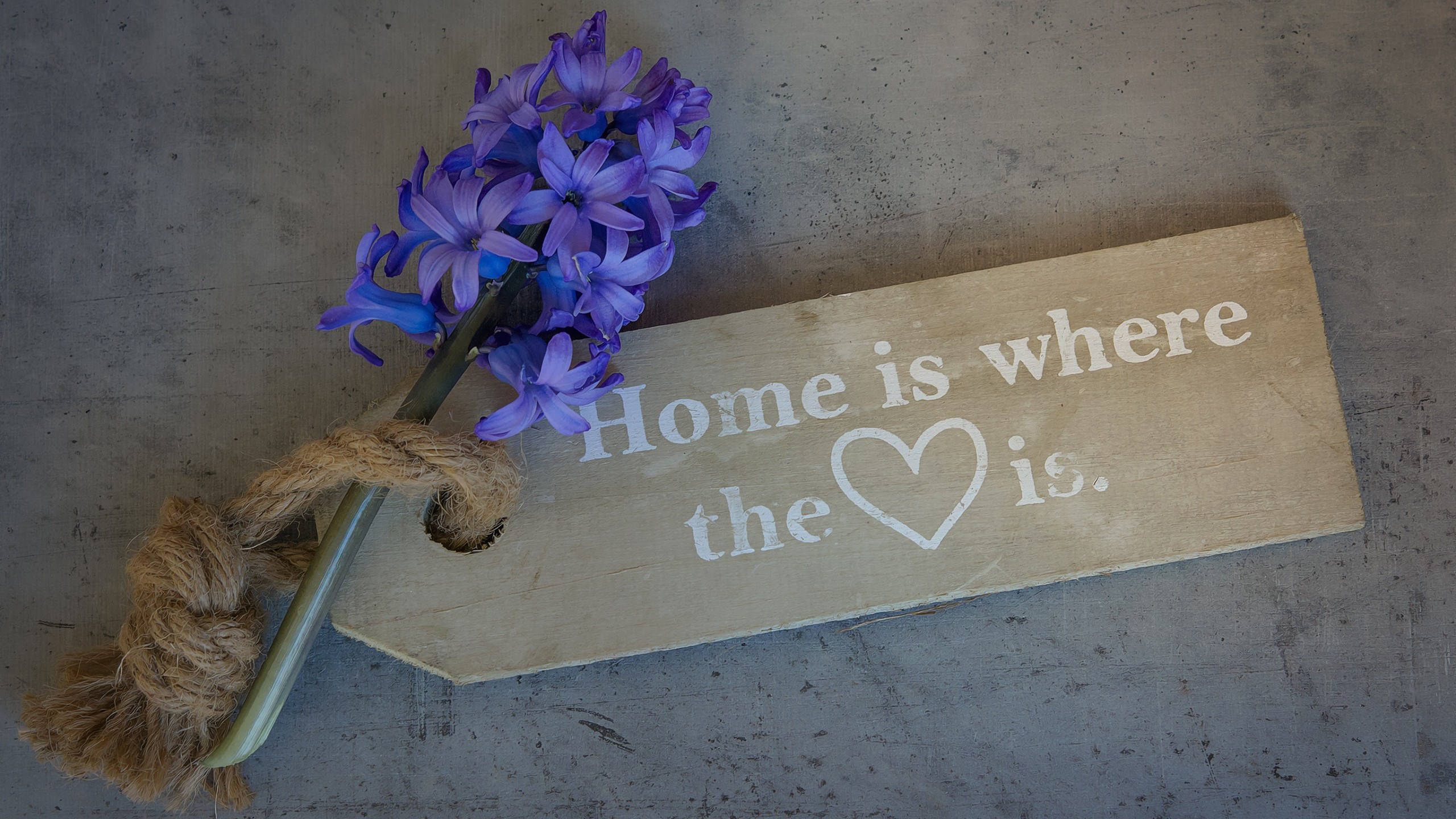 HD inspirational 桌布 壁紙 — 一塊寫有「Home is where the ❤️ is」的質樸木牌，旁邊放著一束用麻繩綁著的紫色風信子，置於灰色混凝土表面上。