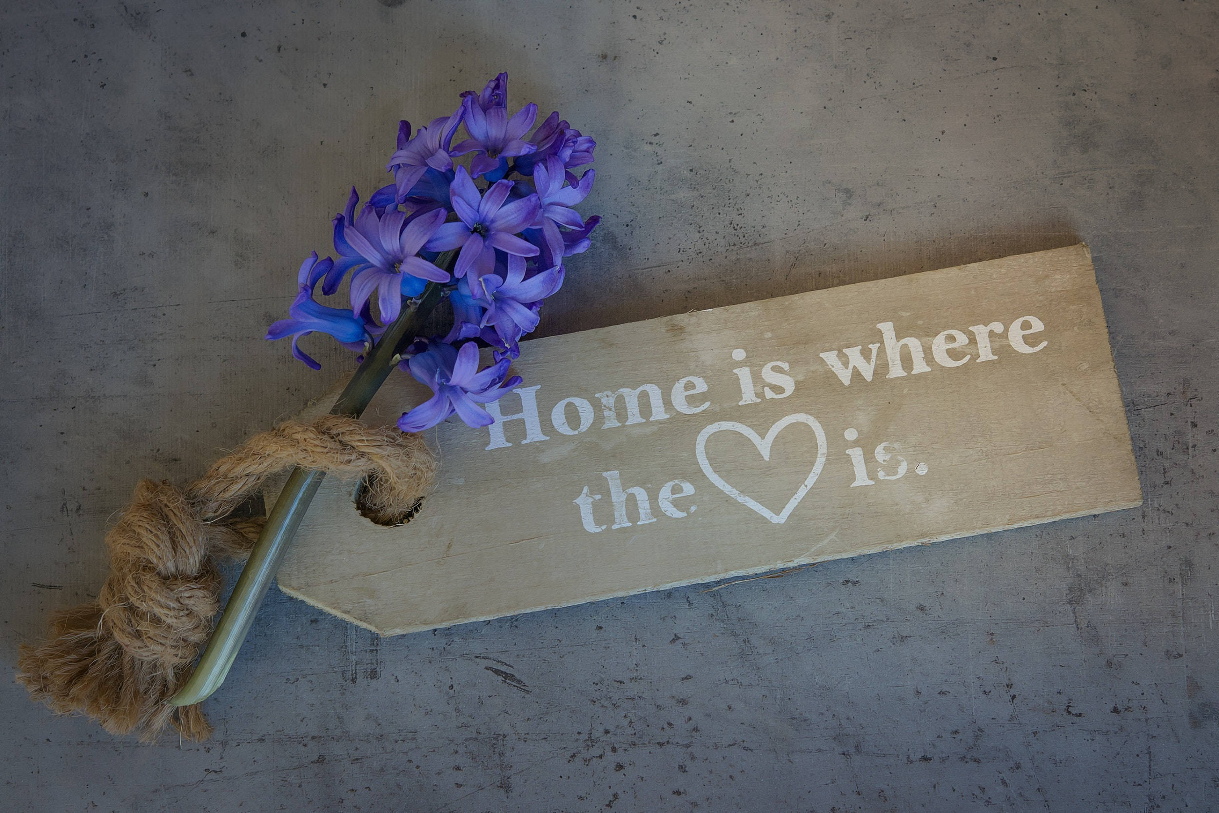 風信子 桌布 壁紙 — 一塊寫有「Home is where the ❤️ is」的質樸木牌，放在混凝土表面上，旁邊是一束用麻繩綁著的紫色風信子。