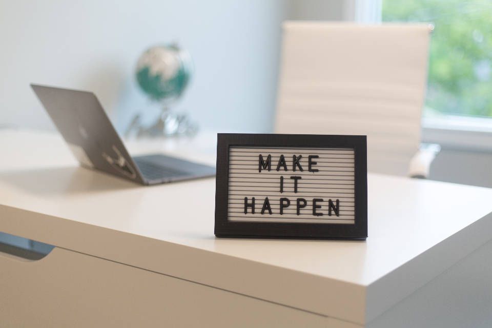 勵志桌面 桌布 壁紙 — 一塊寫有「make it happen」的勵志字母板放在潔白的辦公桌上，旁邊有一台筆記型電腦和一個地球儀，在明亮的辦公室中。