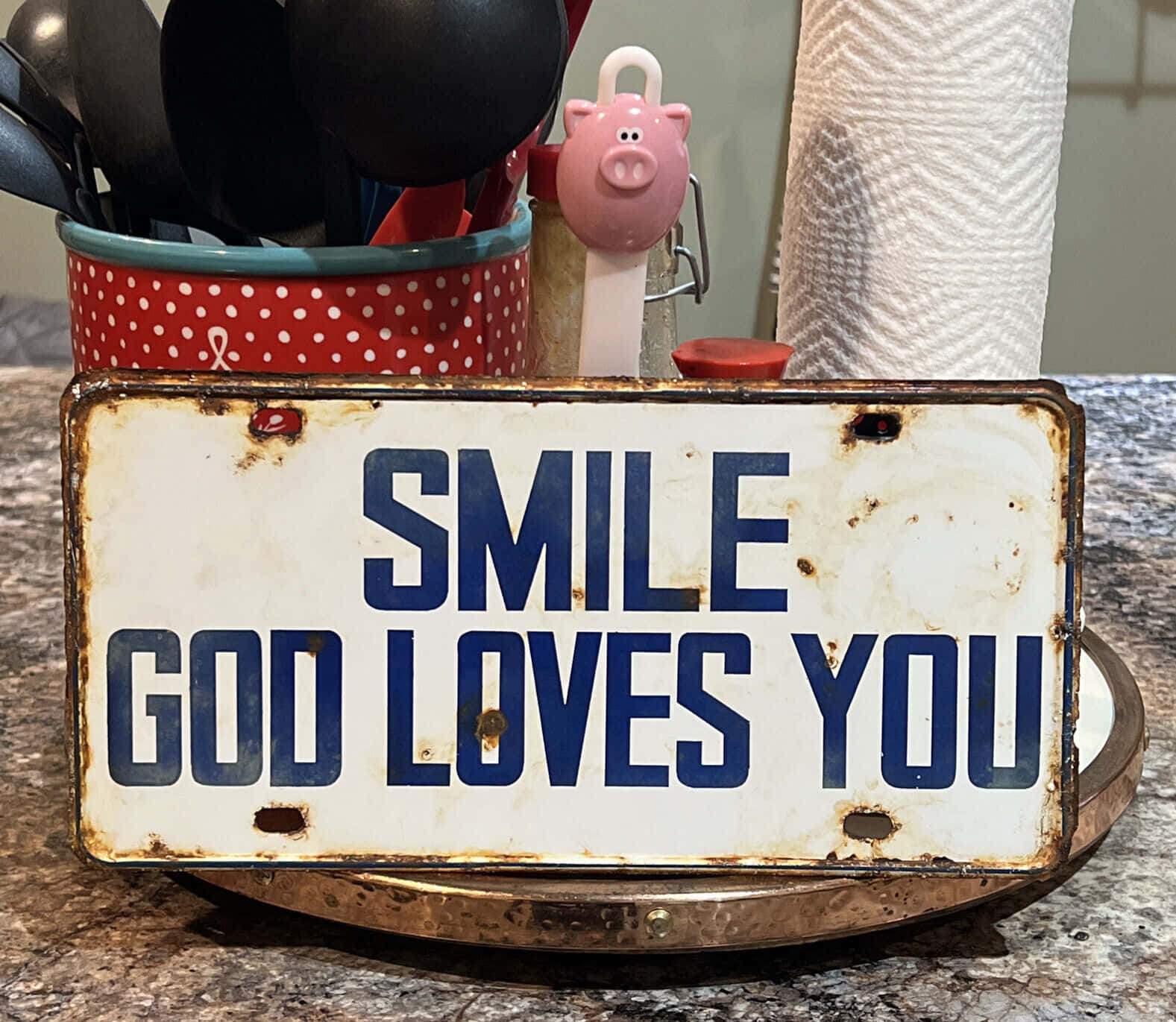 God Loves You 桌布 壁紙 — 一塊寫有「SMILE GOD LOVES YOU」的風化金屬標牌，放在廚房檯面上，旁邊有一個紅色圓點餐具架和一個粉紅色豬形湯匙架。