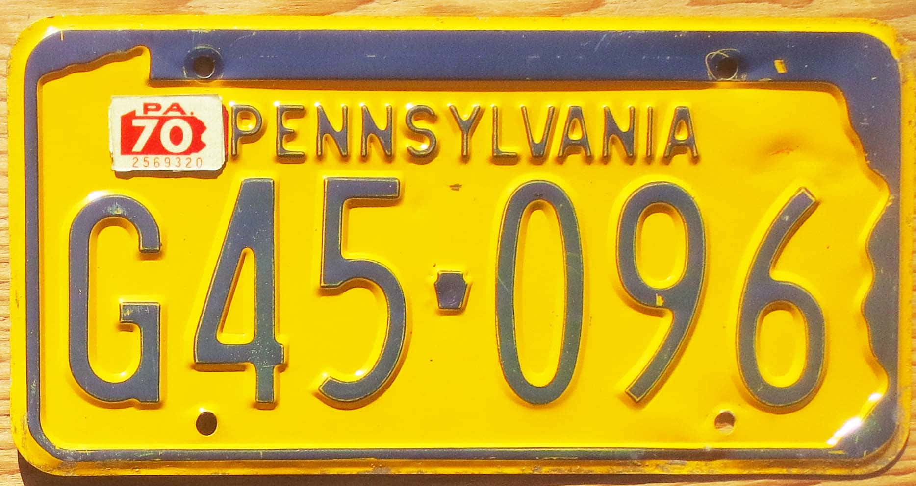 License Plate 桌布 壁紙 — 一塊復古黃色賓夕法尼亞州車牌，帶深藍色邊框和 '70 貼紙，在木紋背景上顯示 G45 096。