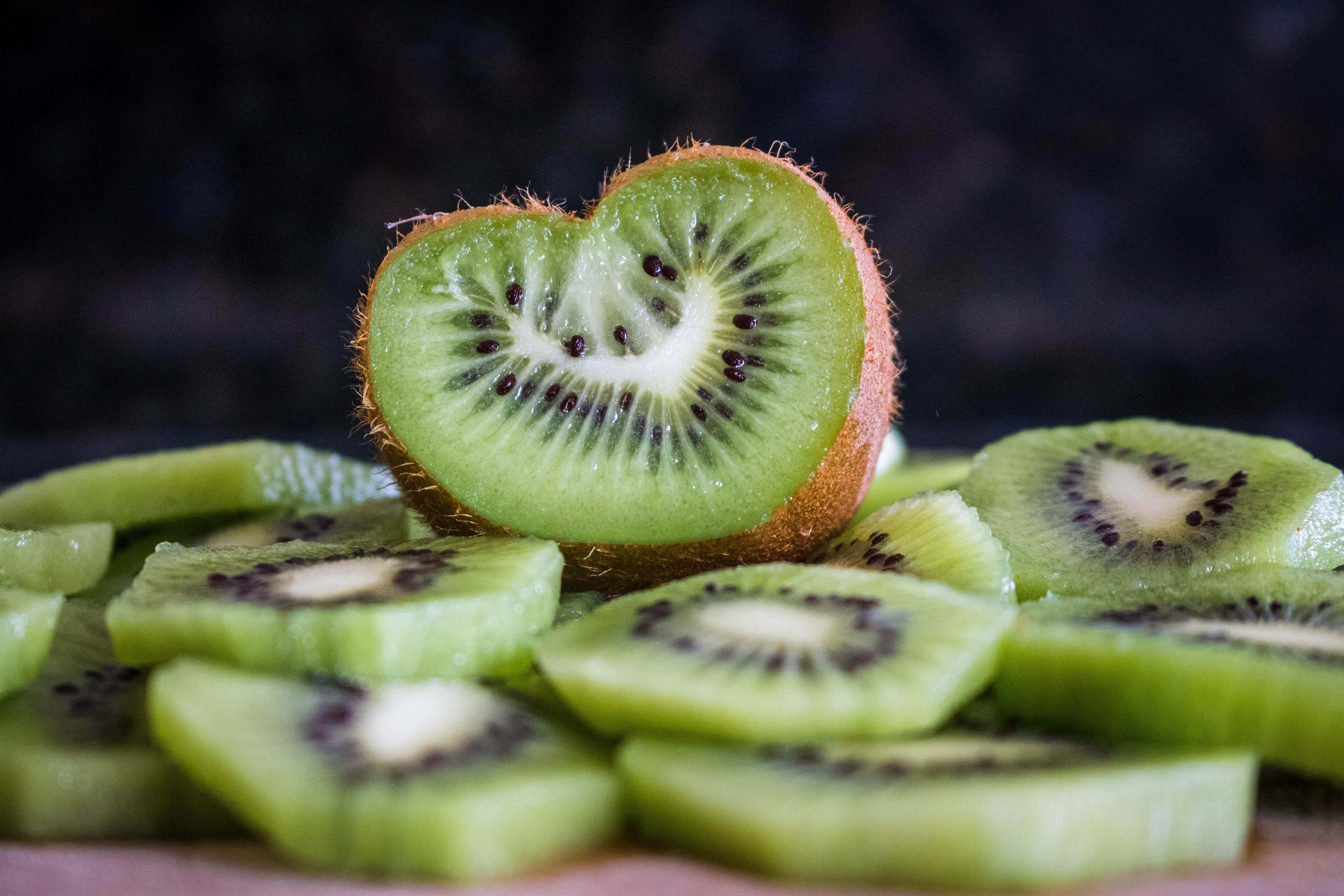Kiwi 桌布 壁紙 — 一塊心形奇異果切片放在一堆新鮮綠色奇異果切片（帶黑色籽）上，背景為深色。