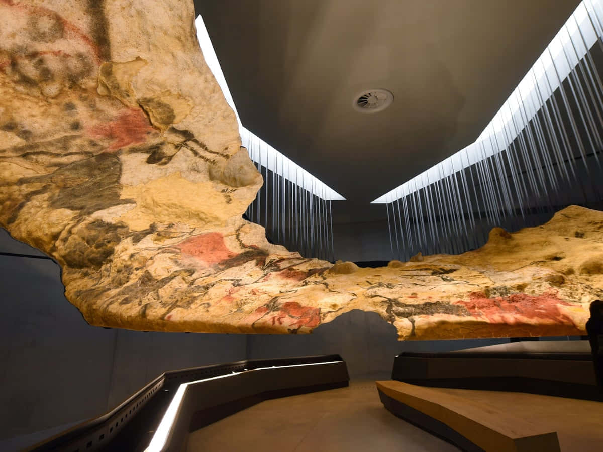 Lascaux Caves 桌布 壁紙 — 一塊懸掛的古代岩石，帶有紅色赭石洞穴壁畫，戲劇性地懸掛在現代博物館中，下方有簡約的燈光和長椅。