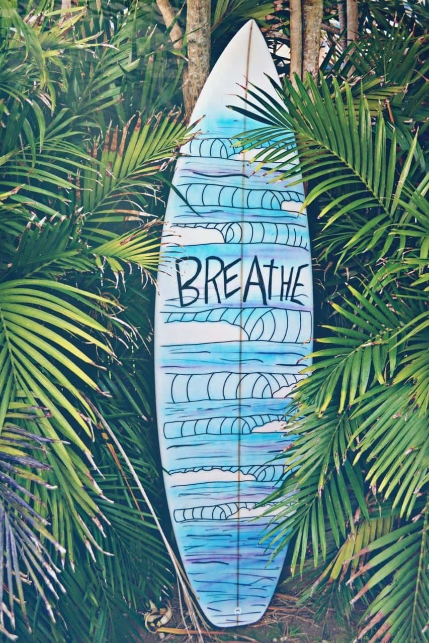 surfing 桌布 壁紙 — 一塊手繪的衝浪板，上面有「BREATHE」字樣和波浪圖案，靠在熱帶環境中茂密的綠色棕櫚葉上。