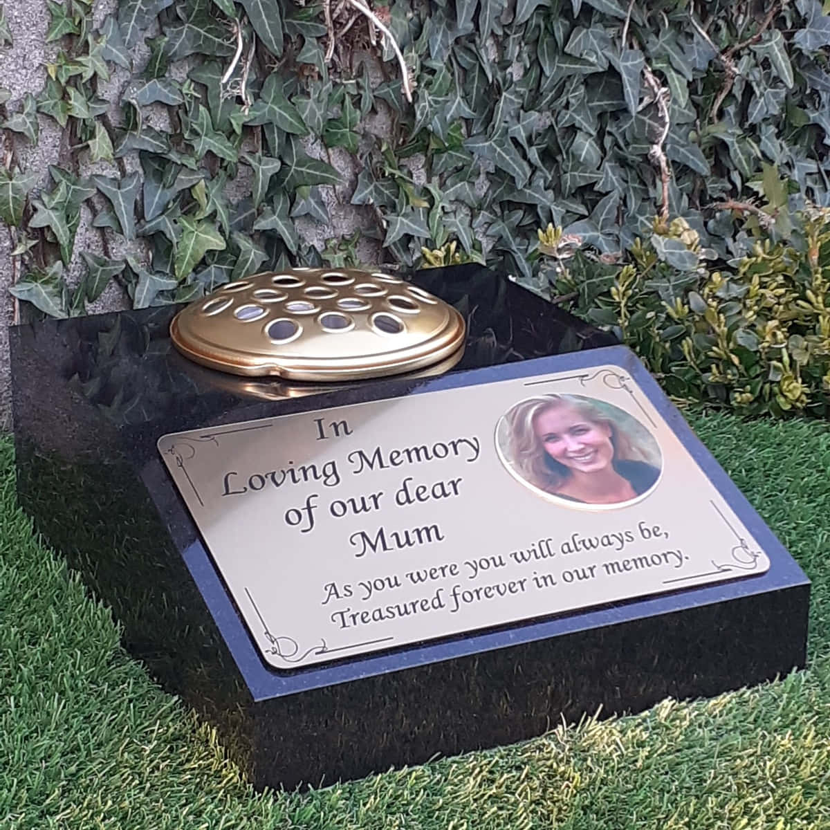 Memorial 桌布 壁紙 — 一塊拋光黑色紀念牌，上面有金色骨灰罈和一位微笑女士的照片，放在草地上，旁邊是常春藤覆蓋的石頭。
