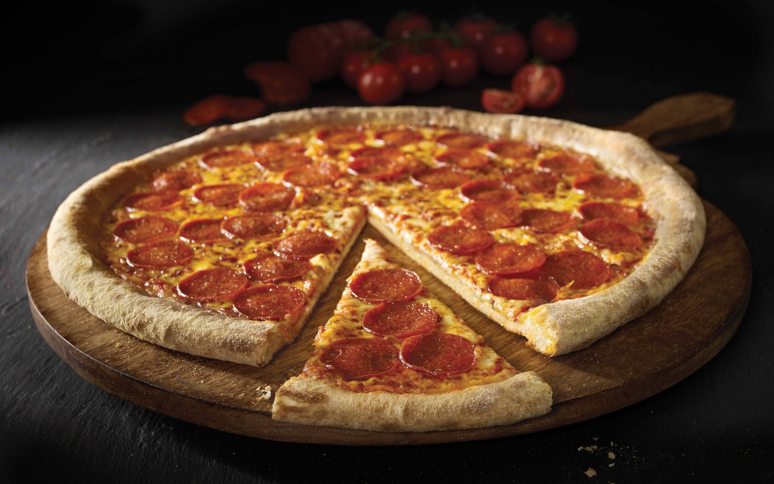 Dominos Pizza 桌布 壁紙 — 一塊新鮮出爐的義大利辣香腸披薩，其中一片被拿走，放在木板上，背景有櫻桃番茄。