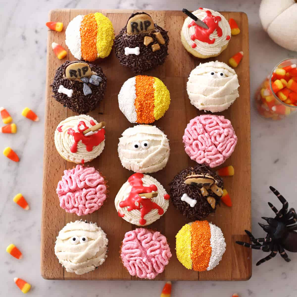 Halloween Cupcakes 桌布 壁紙 — 一塊木板上展示著以木乃伊、大腦和墓碑裝飾的萬聖節主題杯子蛋糕，旁邊有糖果玉米和一隻蜘蛛。