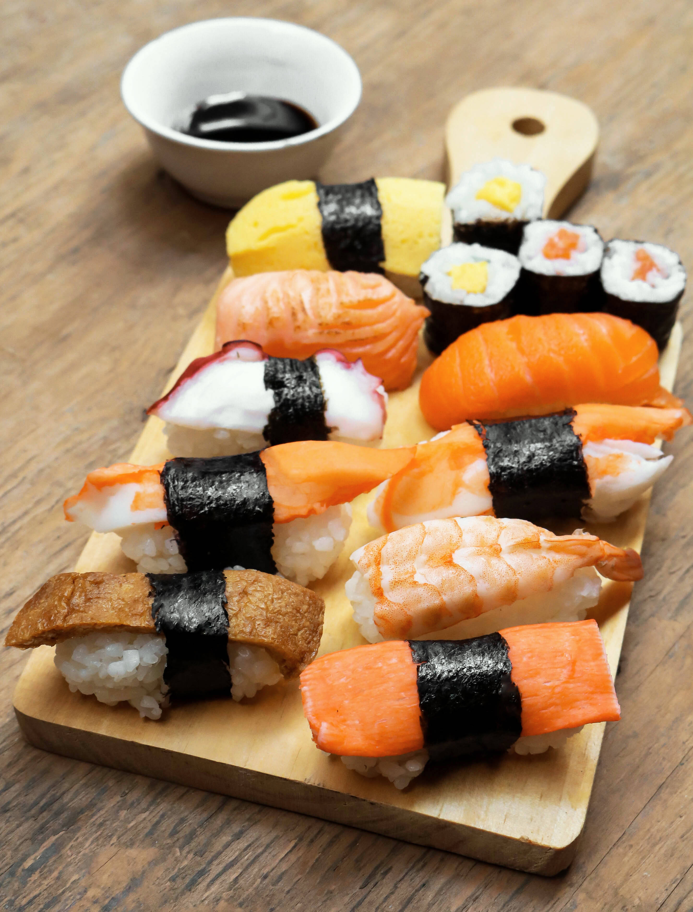 Sushi 桌布 壁紙 — 一塊木板上展示著各種壽司卷和握壽司，包括鮭魚、鮪魚、蝦和玉子，整齊地排列在一個小醬油碗旁邊。