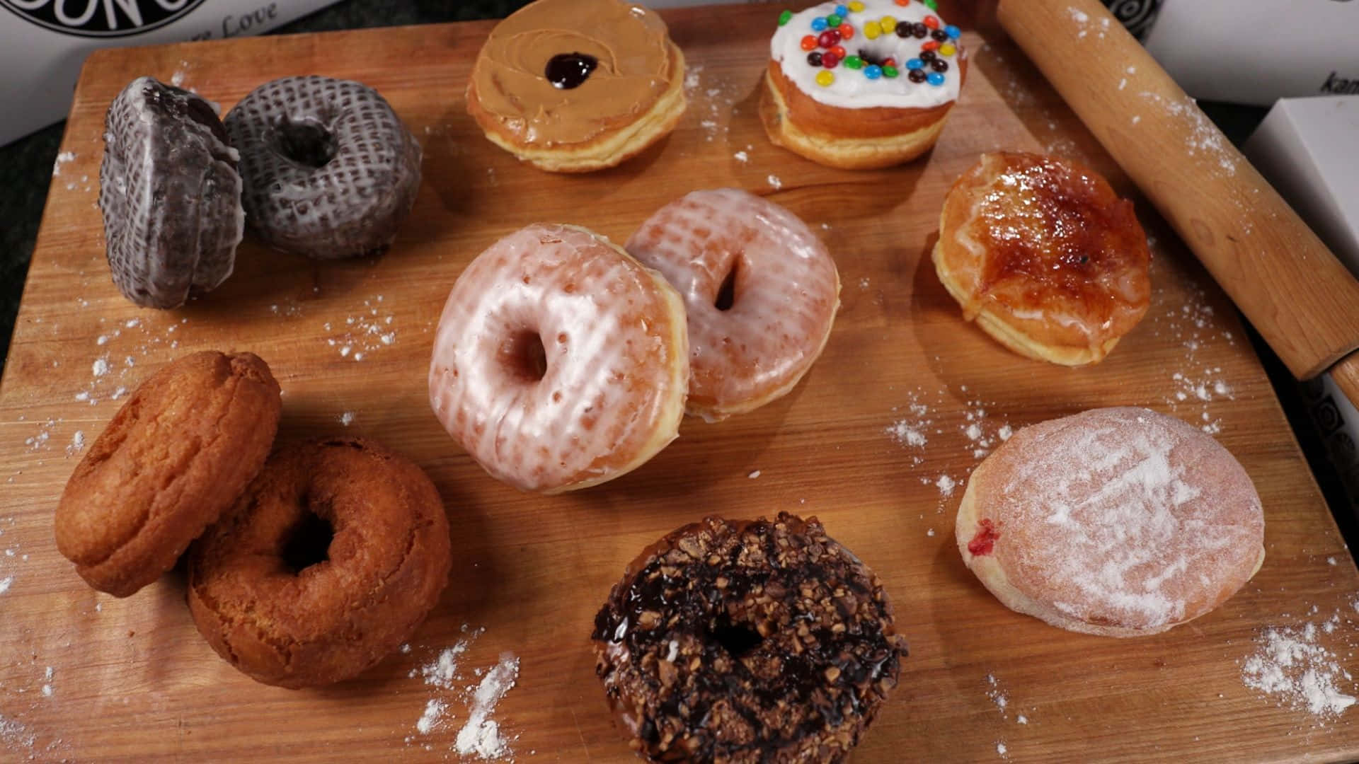 donuts 桌布 壁紙 — 一塊木板上展示著各種帶有糖霜、彩針和糖粉的甜甜圈，旁邊還有一個擀麵棍。