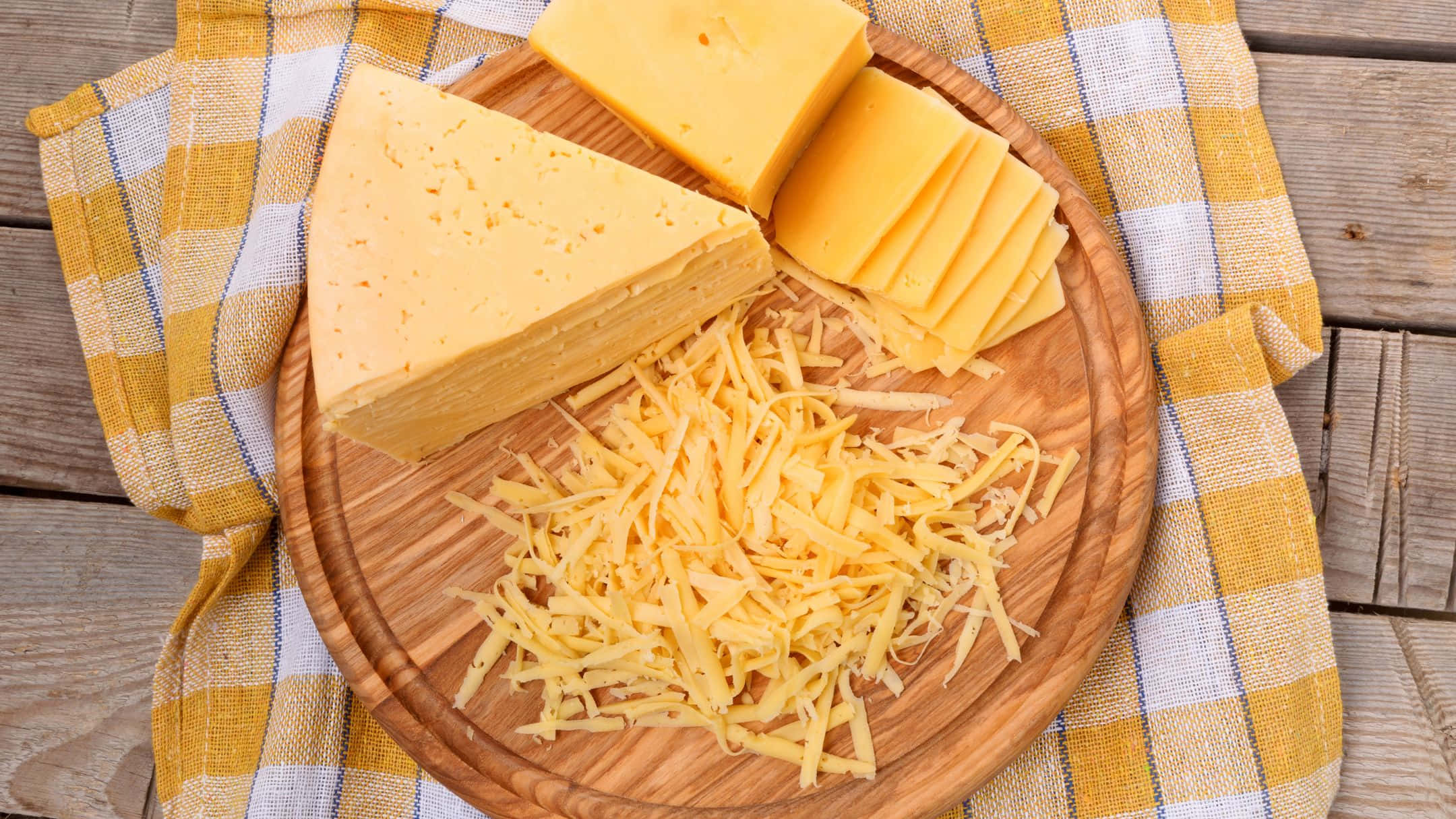 Cheese 桌布 壁紙 — 一塊木板上放著一塊楔形切片起司和碎起司，放在黃色格子布上，置於鄉村風格的木桌上。