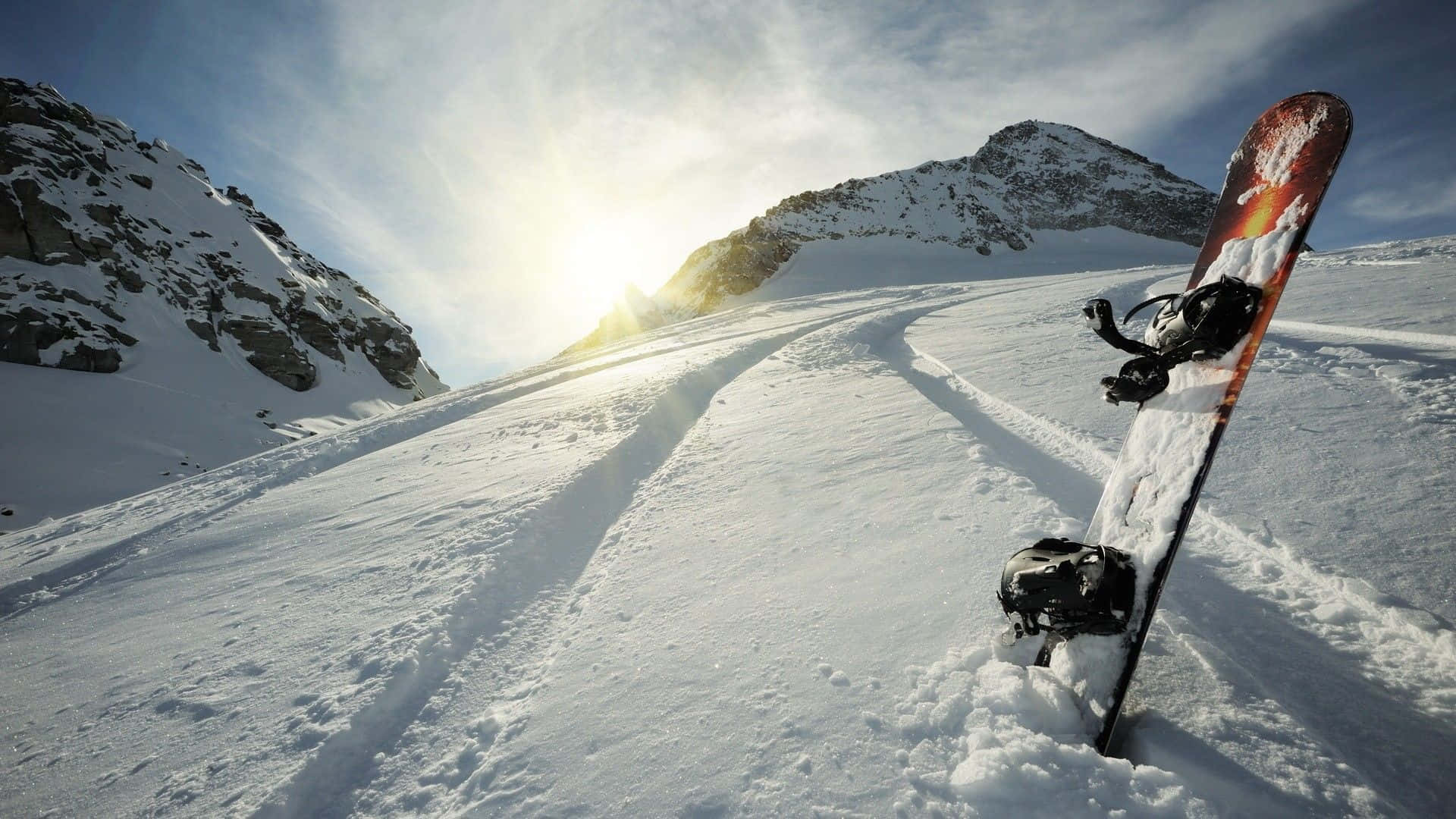 Winter Sports 桌布 壁紙 — 一塊滑雪板直立在新雪粉上，位於陽光明媚的山坡，有蜿蜒的軌跡通往遠方山峰。