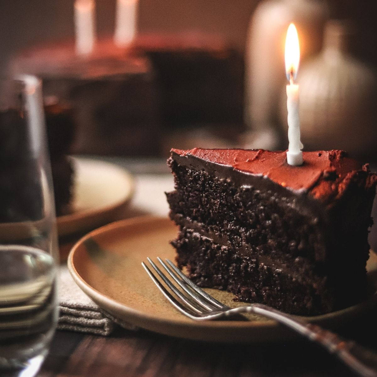 Birthday Cake 桌布 壁紙 — 一塊濃郁的巧克力蛋糕，上面插著一根點燃的蠟燭，放在盤子上，旁邊有叉子和玻璃杯，在溫暖昏暗的燈光下。