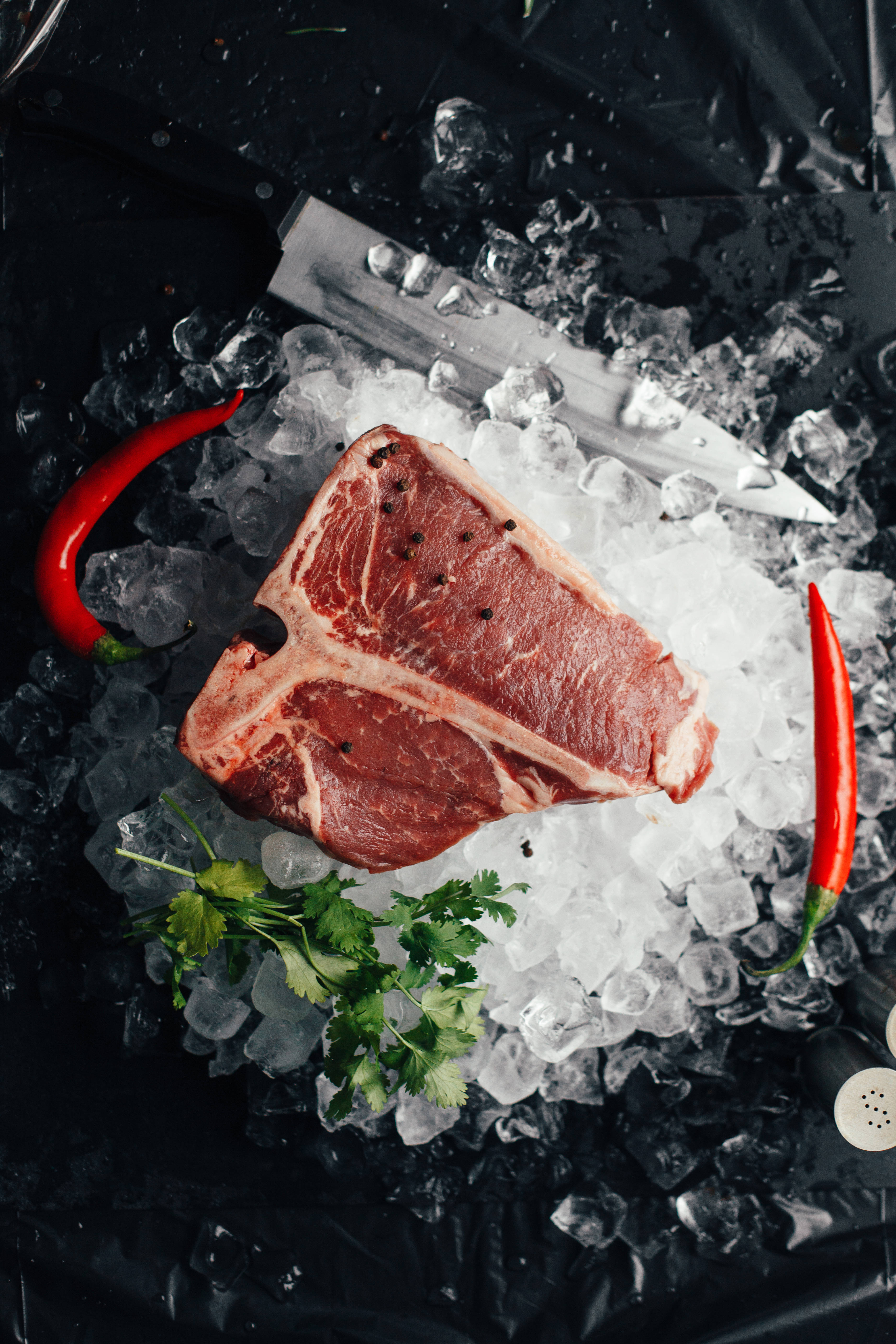 meat 桌布 壁紙 — 一塊生T骨牛排放在碎冰上，配上紅辣椒和香菜，附近有一把鋒利的刀在深色表面上。