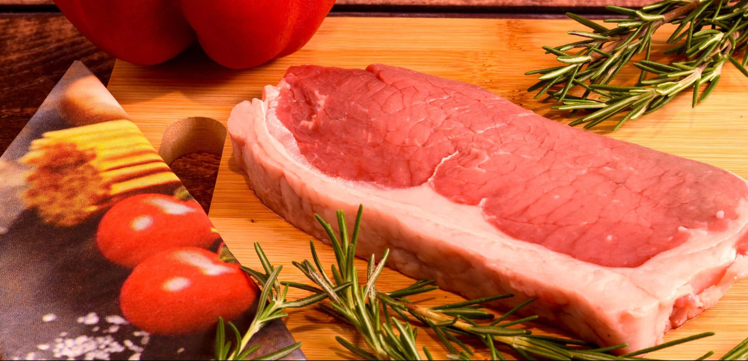 Red Meat 桌布 壁紙 — 一塊生牛肉排放在木板上，周圍有新鮮迷迭香、小番茄和紅甜椒。