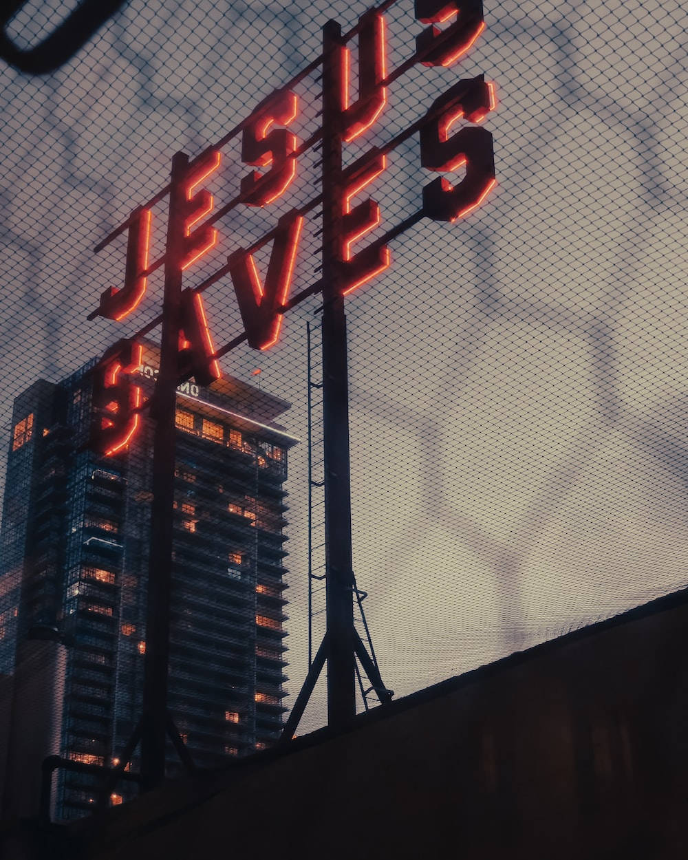 手機 桌布 壁紙 — 一塊發光的紅色霓虹燈牌，上面寫著「JESUS SAVES」，豎立在鐵絲網圍欄後面，背景是黃昏時分亮燈的摩天大樓。