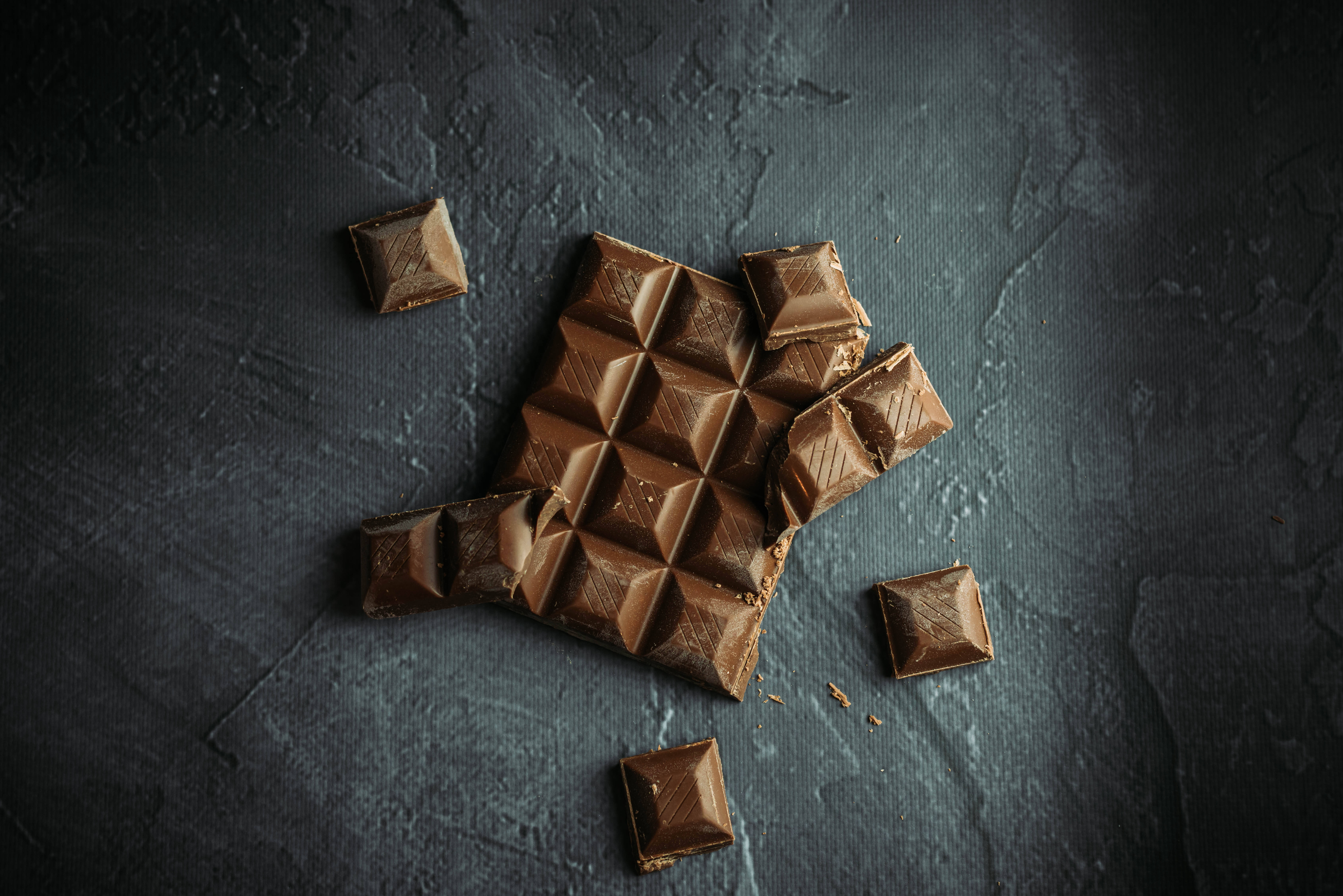Chocolate 桌布 壁紙 — 一塊破碎的巧克力條，散落著方塊，放在深色紋理表面上，在戲劇性的燈光下。