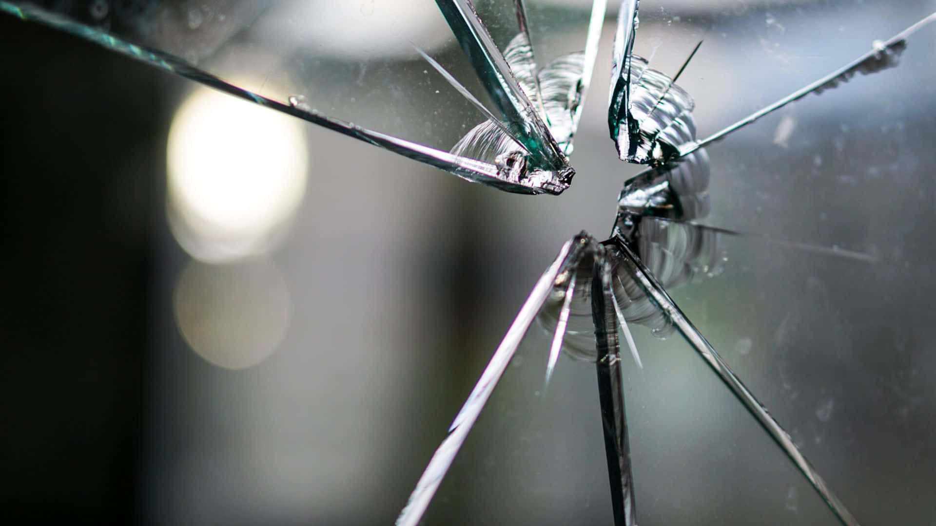 Broken Glass 桌布 壁紙 — 一塊破碎的玻璃板，從中央撞擊點放射出尖銳裂紋，背景模糊。