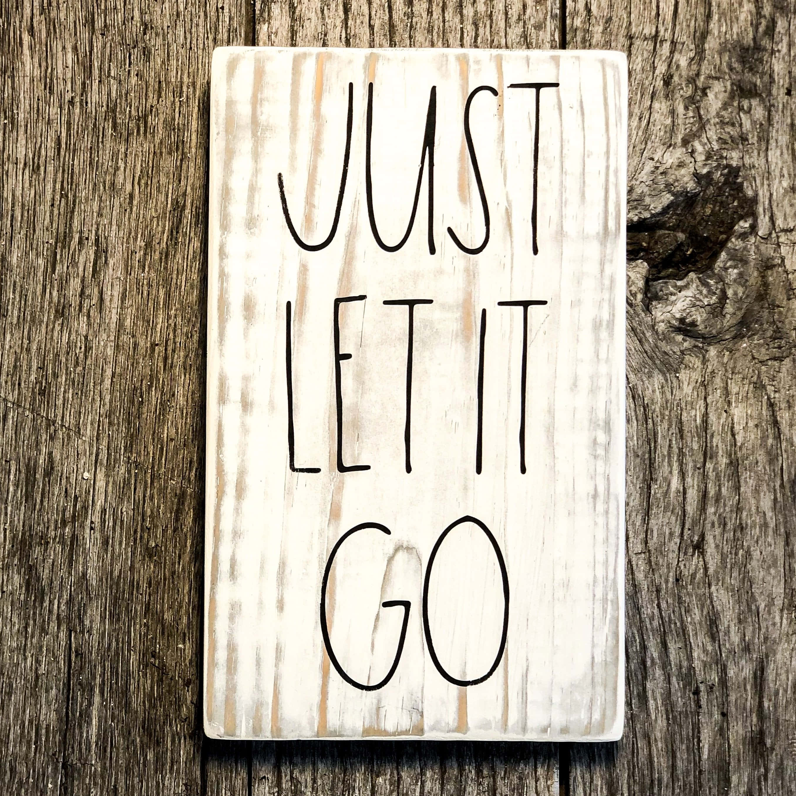 let it go 桌布 壁紙 — 一塊經風化的白色木牌，上面有黑色手寫文字「JUST LET IT GO」，擱在粗糙有紋理的木質表面上。