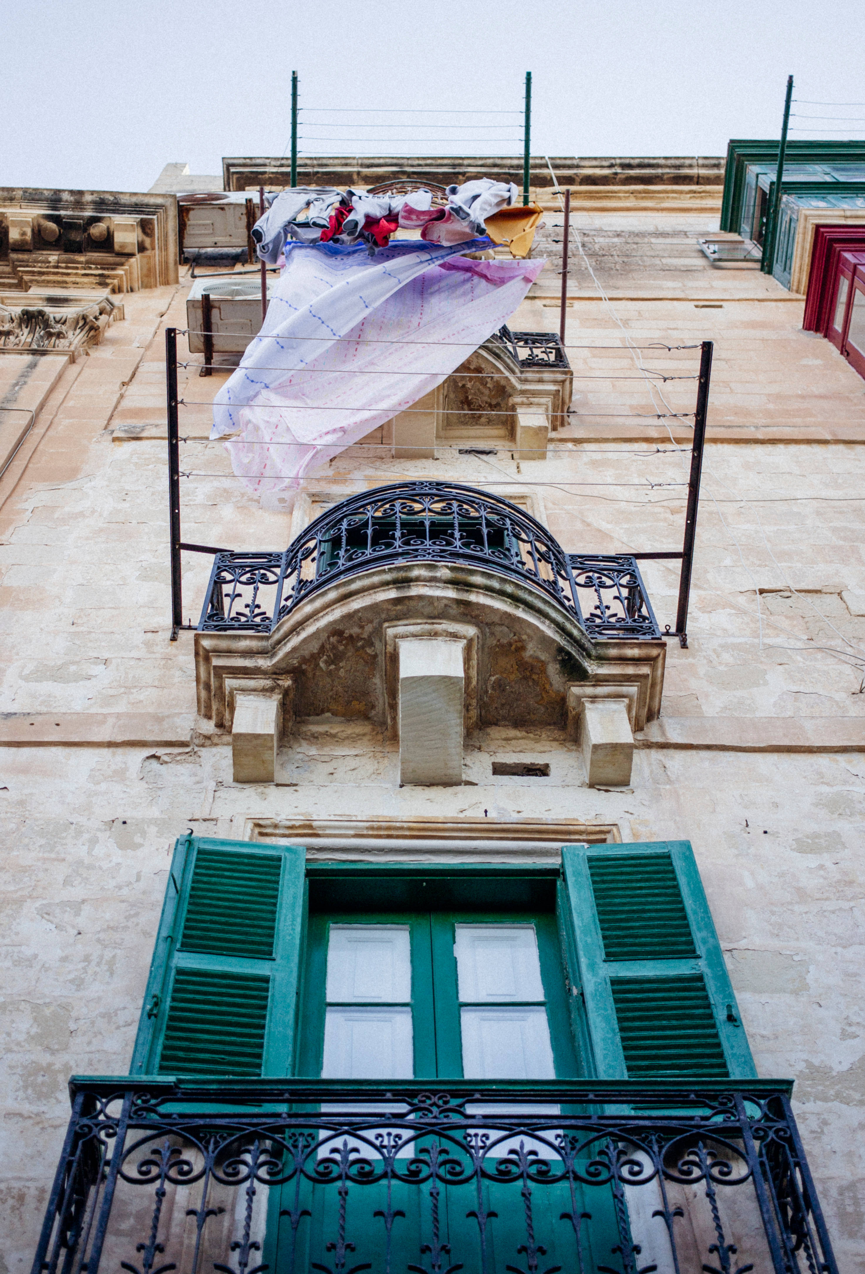Malta 桌布 壁紙 — 一塊色彩繽紛的大床單在風中戲劇性地飄揚，位於一座風化的石建築頂部，建築帶有華麗的綠色百葉窗和黑色鐵藝陽台。