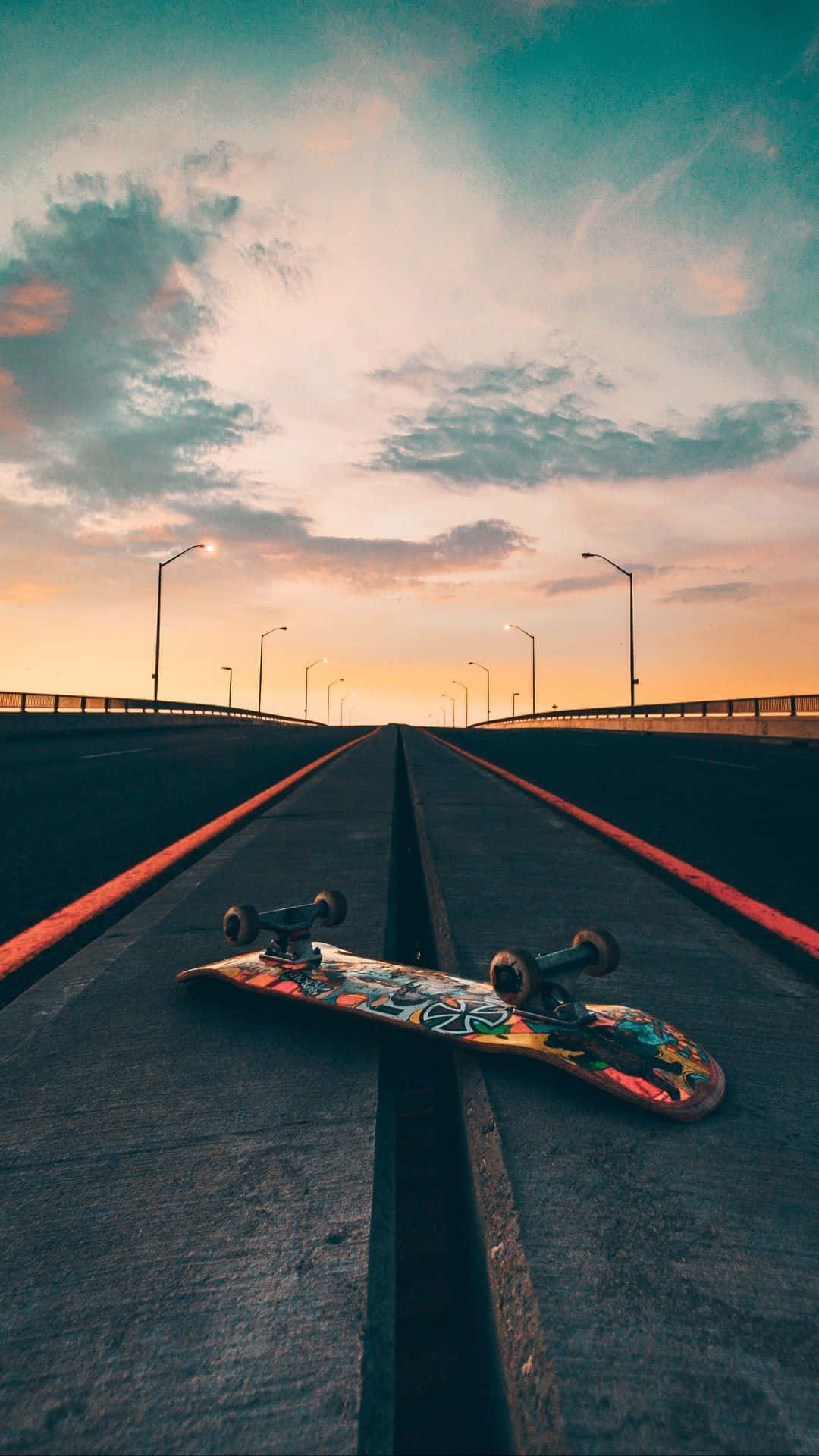 Retro Skateboard 桌布 壁紙 — 一塊色彩繽紛的滑板在日落時分放在橋上，上方是溫暖的橙色和青綠色的天空。