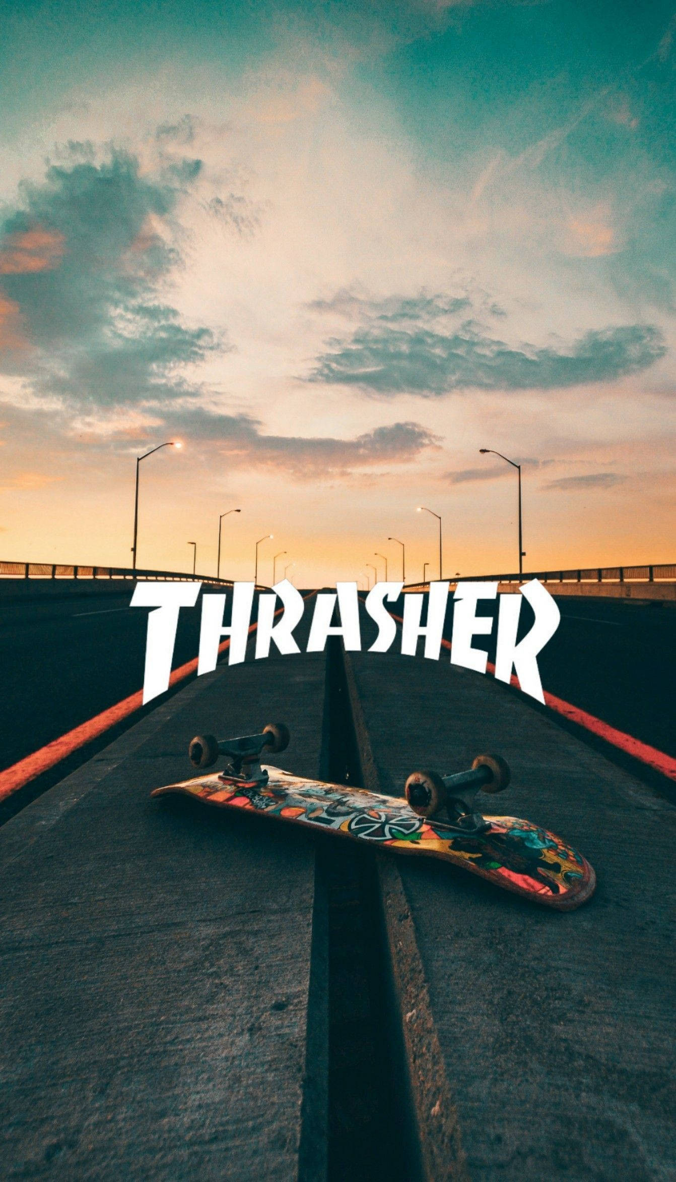 Thrasher 桌布 壁紙 — 一塊色彩繽紛的滑板放在混凝土橋上，夕陽西下，天空中有橙色和藍綠色的雲朵，THRASHER 字樣以粗體白色字母呈現。