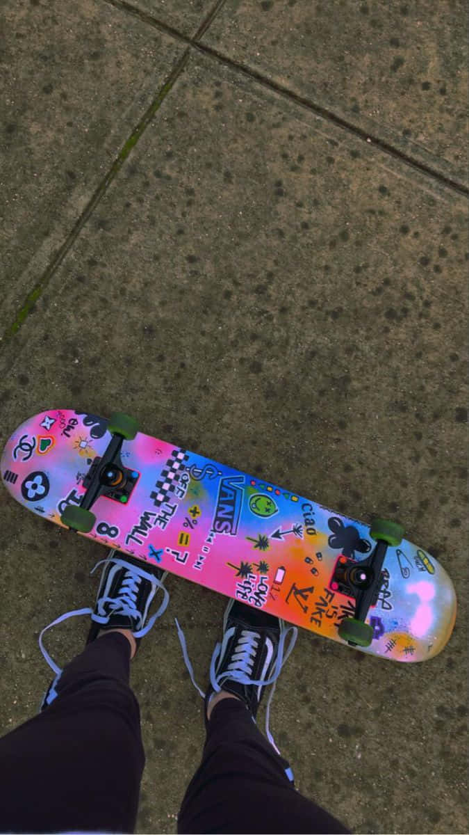 Retro Skateboard 桌布 壁紙 — 一塊色彩繽紛貼紙裝飾的滑板放在混凝土人行道上，由一位穿黑色運動鞋的人從上方觀看。