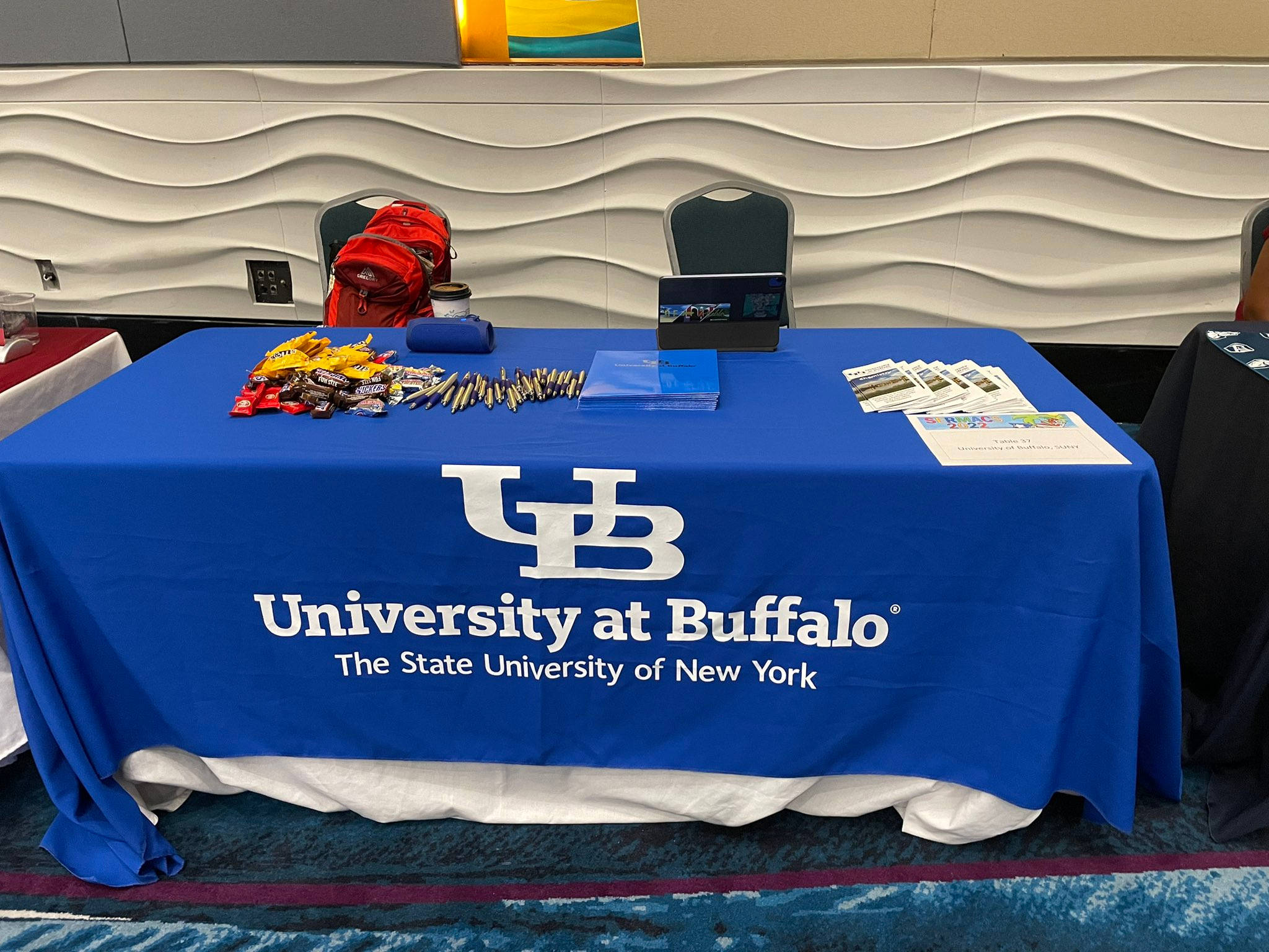 學校 桌布 壁紙 — 一塊藍色的University at Buffalo桌布覆蓋著一張擺有宣傳物品、零食、筆和宣傳單張的活動桌。
